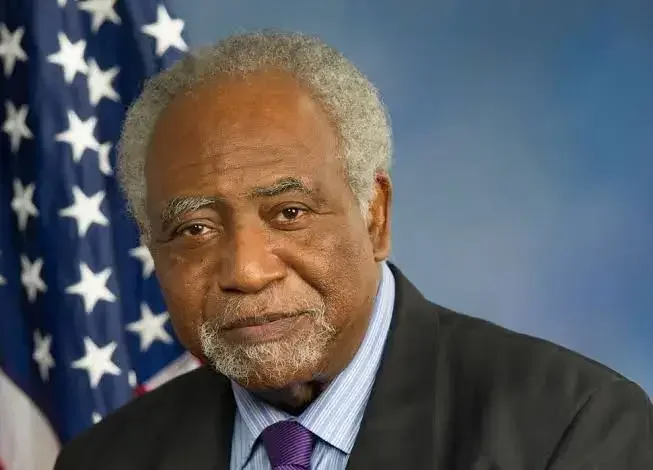 A portrait of an elderly African American man with gray hair and beard, dressed in a suit and tie, standing in front of an American flag.