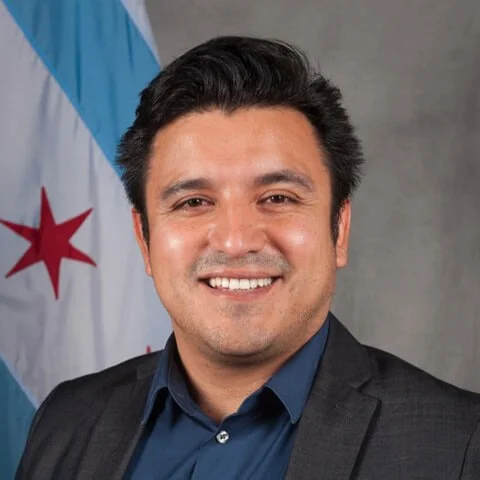 A smiling young Hispanic man in a dark suit and blue shirt standing in front of a gray background and a flag with a red star and blue stripes.