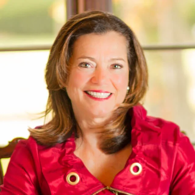 A woman with shoulder-length brown hair wearing a red blouse, smiling while seated indoors.