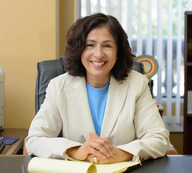 A woman with short dark curly hair smiling and sitting at a desk in an office, wearing a cream blazer and blue shirt.