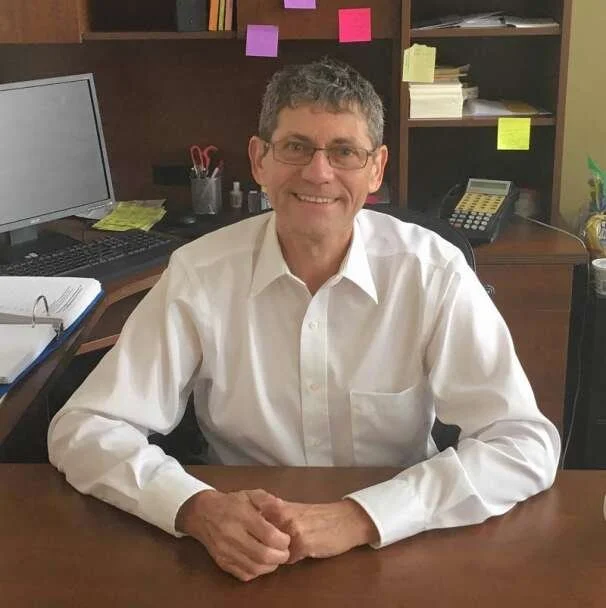 A man with glasses and gray hair smiling, sitting at a desk in an office with books and sticky notes on shelves behind him.