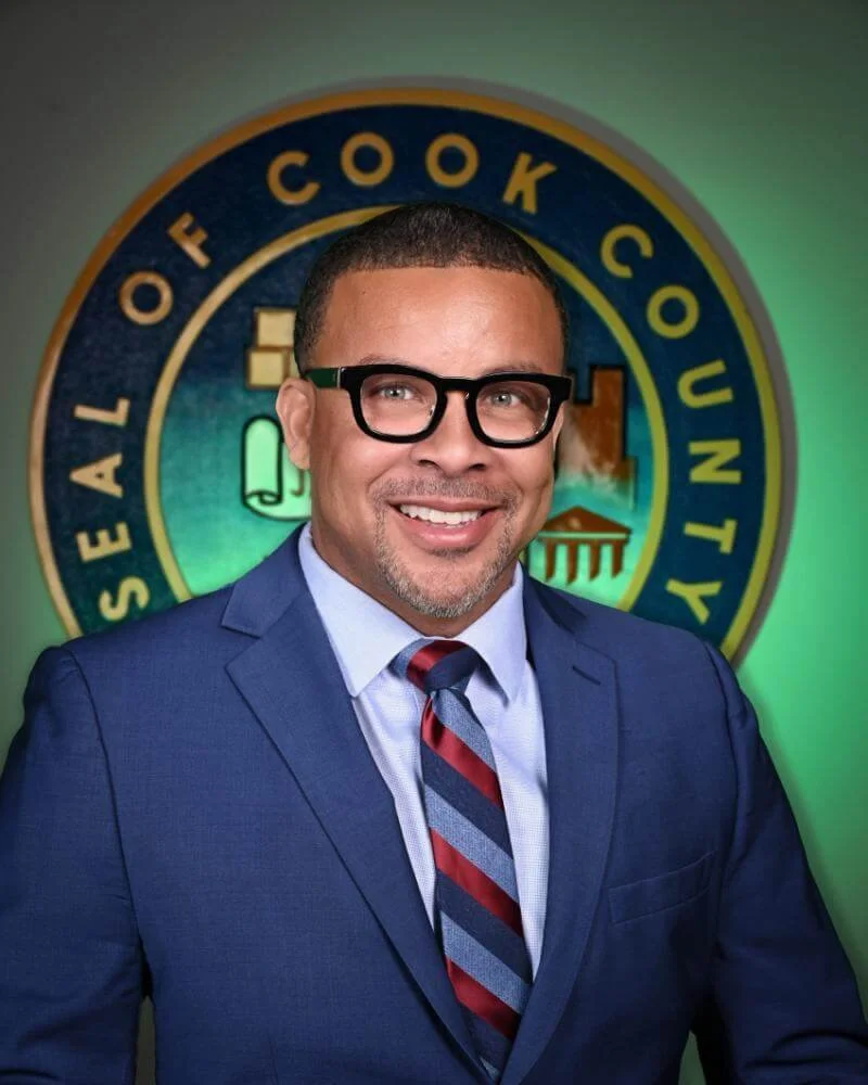An African American man in a blue suit, light blue shirt, and striped tie standing in front of a Seal of the Office of Cook County, Illinois, smiling at the camera.