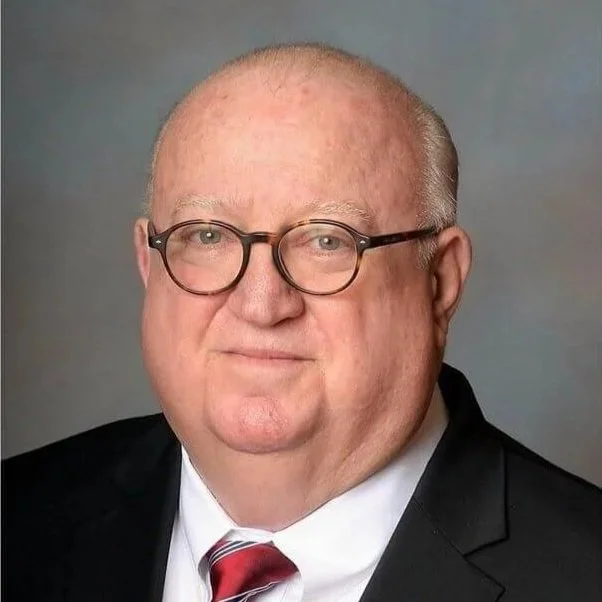 A middle-aged man with glasses in a professional portrait. He has a bald head, light skin, and is wearing a black suit, white shirt, and red striped tie.