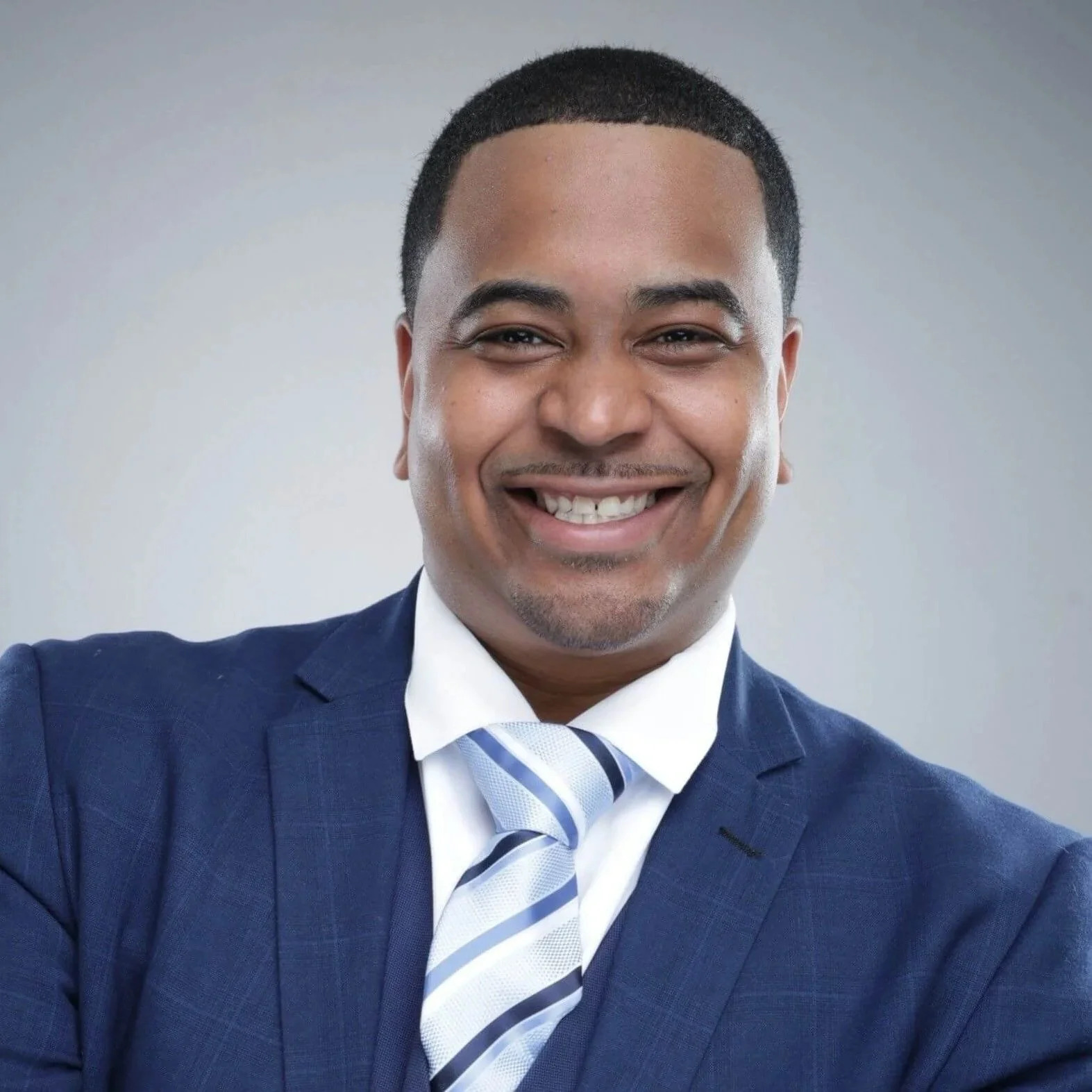 Smiling African American man in a dark blue suit, light blue and white striped tie, white shirt, with short black hair, posing confidently against a plain gray background.
