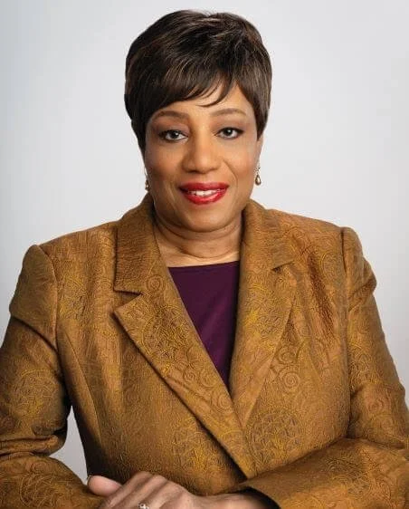An African American woman with short dark hair wearing a gold blazer over a purple top, smiling against a plain light gray background.