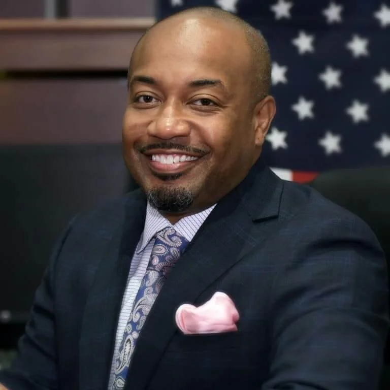 A smiling African American man in a dark suit with a patterned tie and pink pocket square, seated in front of a blurred American flag background.