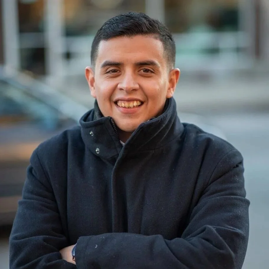 A young Hispanic man smiling with arms crossed, wearing a black jacket, outdoors in an urban setting.