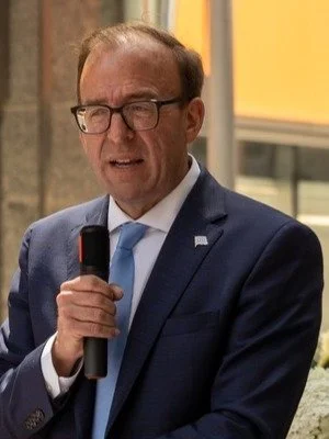 A man wearing glasses in a dark suit and blue tie speaking into a microphone at an outdoor event.