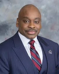 An African American man wearing a navy suit, white shirt, and red striped tie, standing against a textured blue-gray background.