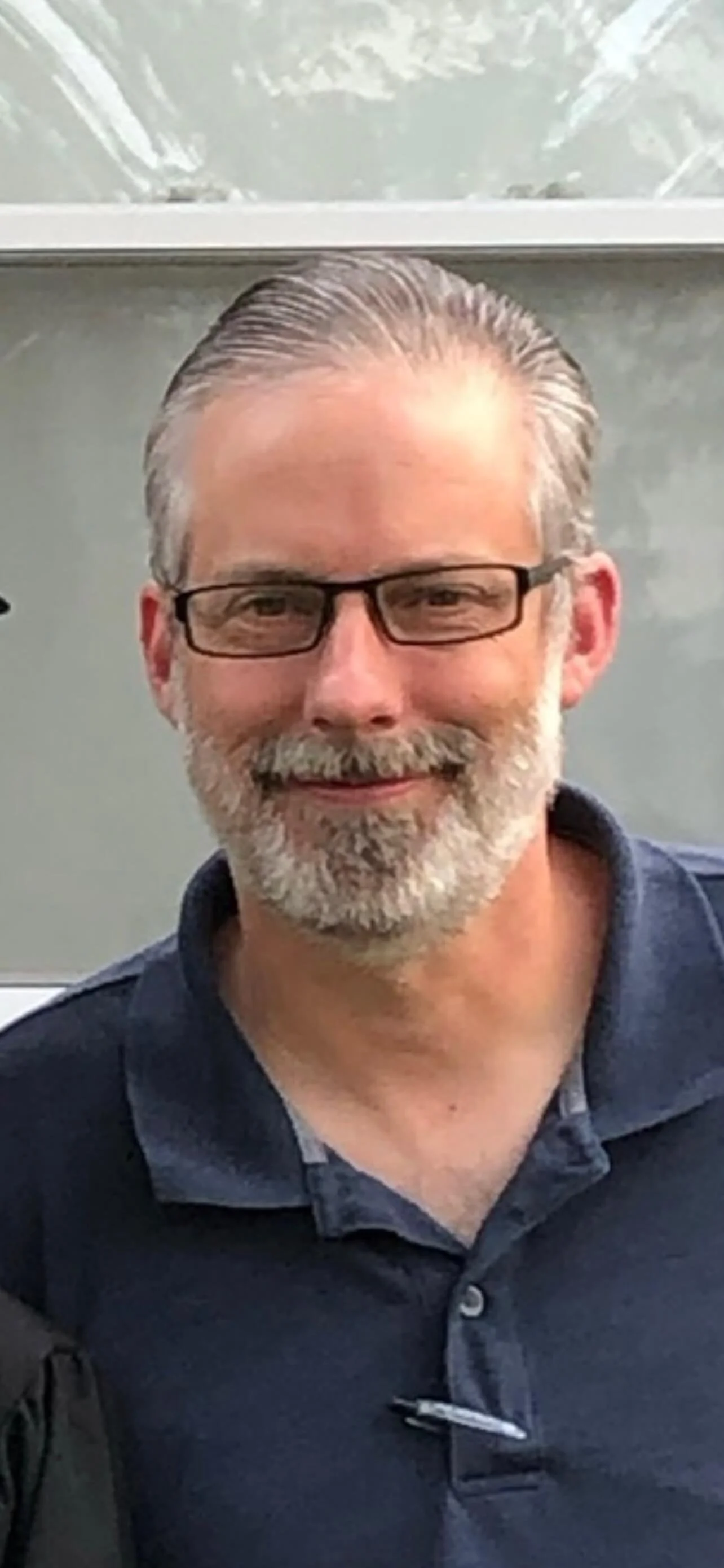 A middle-aged man with gray hair, glasses, and a beard smiling at the camera, wearing a dark polo shirt, standing outdoors.