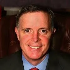 A middle aged man smiling, wearing a blue shirt and a dark blazer.