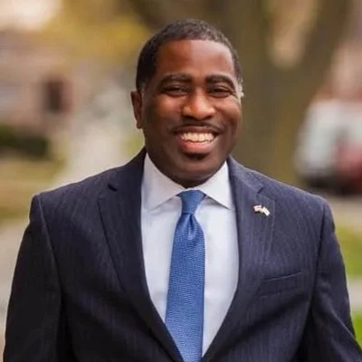 A smiling man in a dark suit and blue tie standing outdoors.