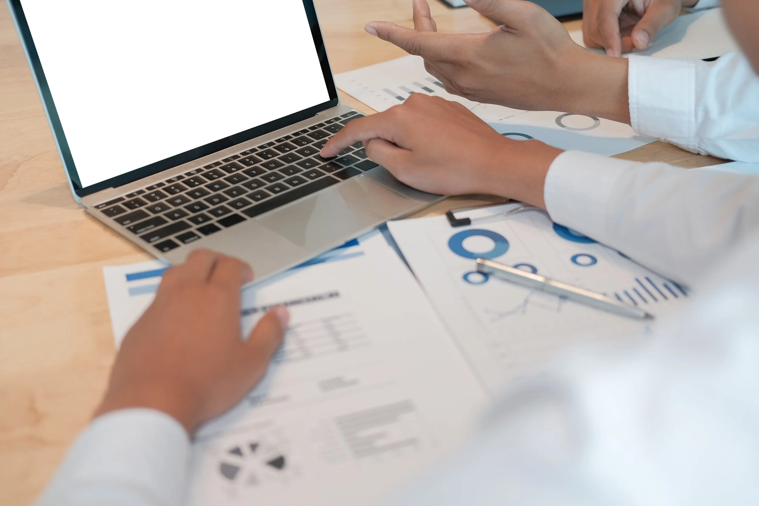 Two people working on a laptop and discussing documents with charts and graphs on a wooden table.