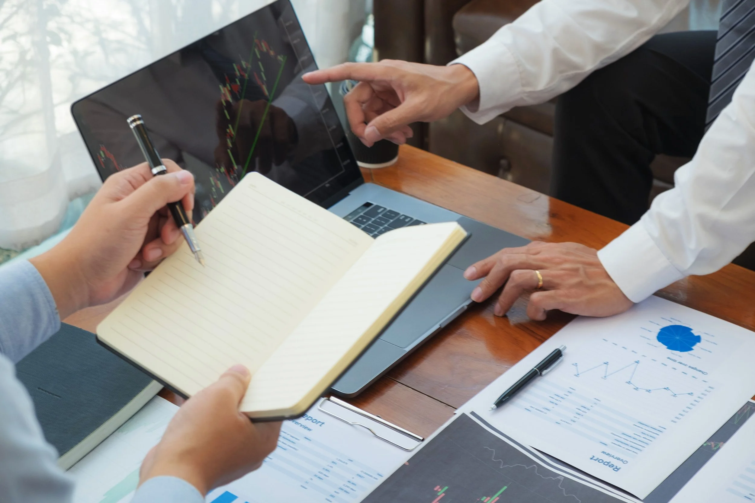 Two people discussing financial data and stock charts. One person is holding a notebook and pen, the other is pointing at a laptop screen showing stock charts. Documents with graphs and reports are spread on the table.