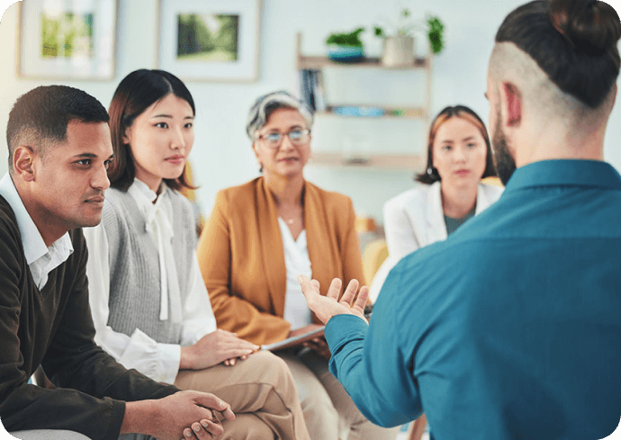 A diverse group of five people engaged in a discussion in a casual office or therapy setting, with a man in a blue shirt speaking and a woman with glasses and gray hair listening.