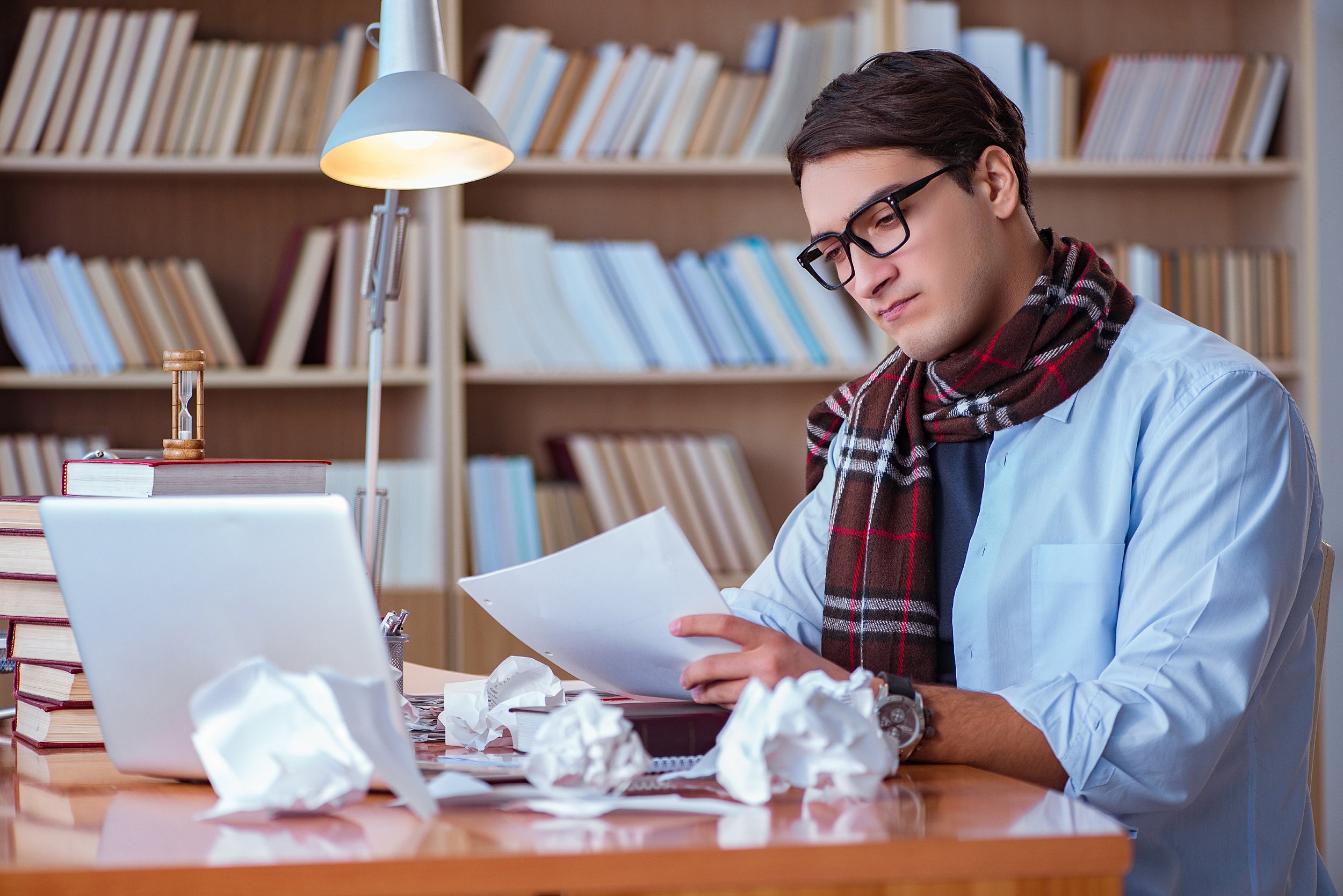 A young man with glasses, wearing a light blue shirt and a plaid scarf, sitting at a cluttered desk, reading a piece of paper, with crumpled papers and a laptop in front of him. A bookshelf filled with books is in the background.
