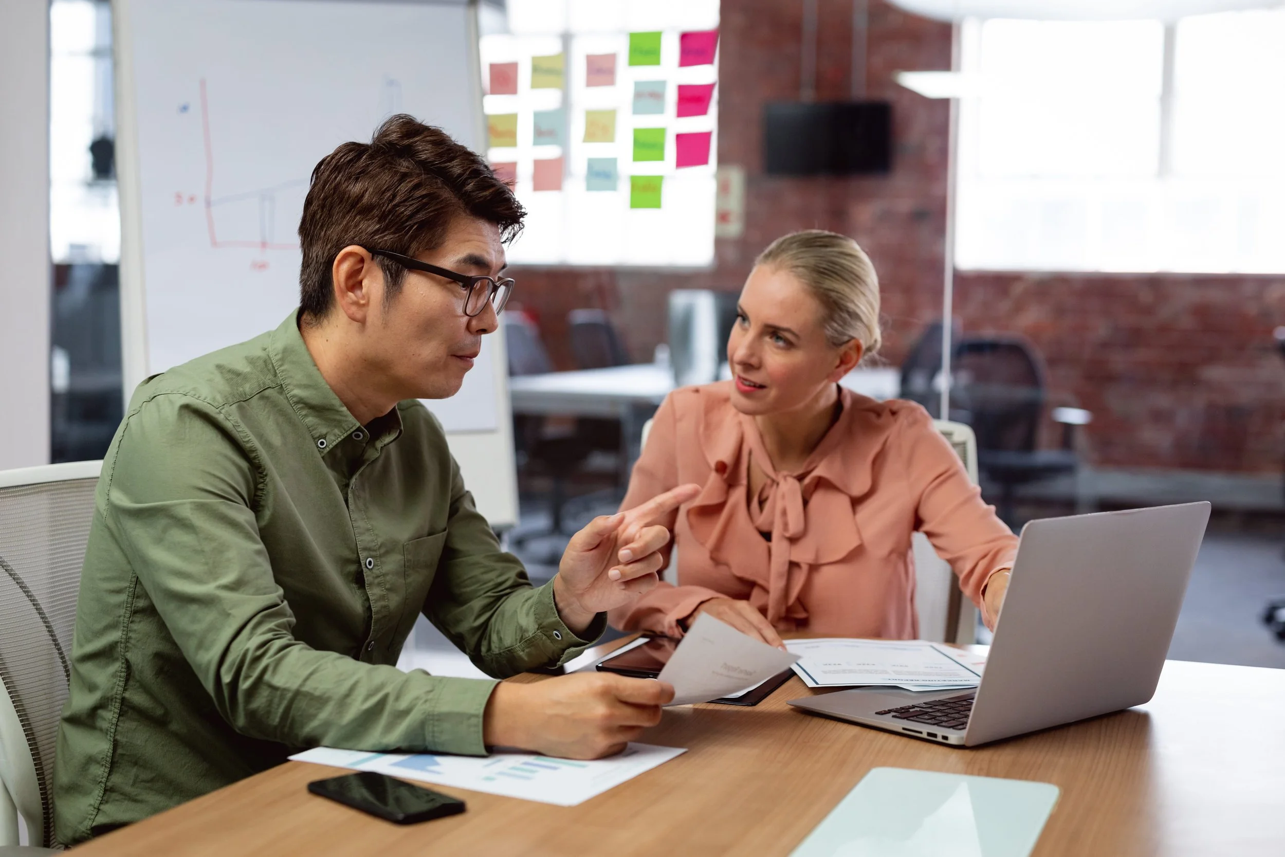 Two colleagues having a discussion at a desk in an office, with a whiteboard and colorful sticky notes in the background.