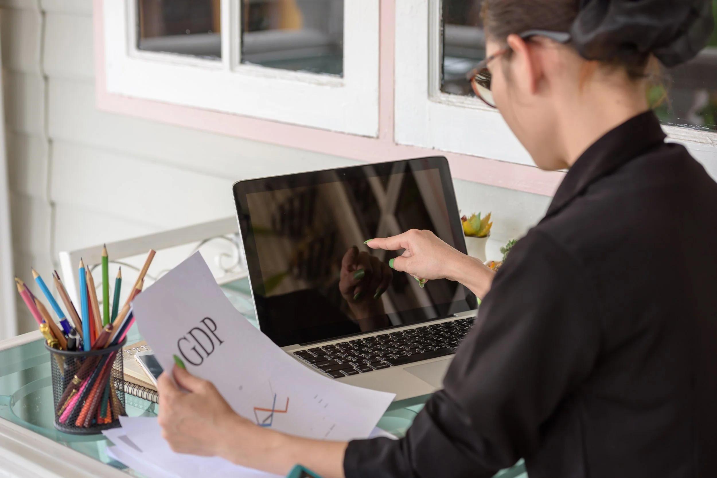 A woman sitting at a desk working on a laptop, holding a paper labeled 'GDP', with colored pencils in a container nearby.
