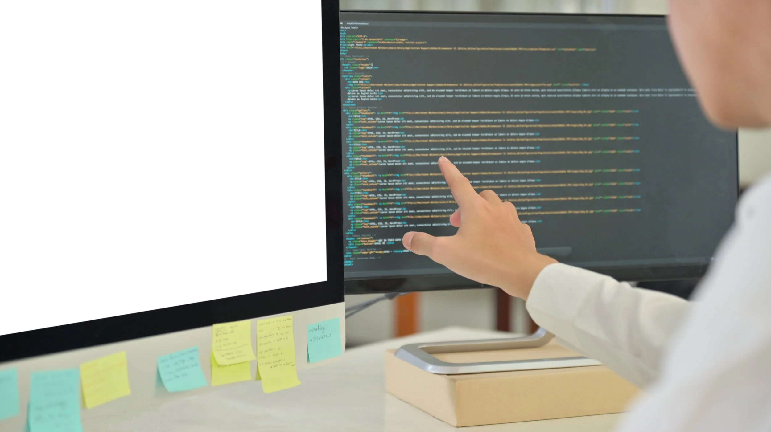 A person pointing at lines of code on a computer monitor, with sticky notes on the desk below.