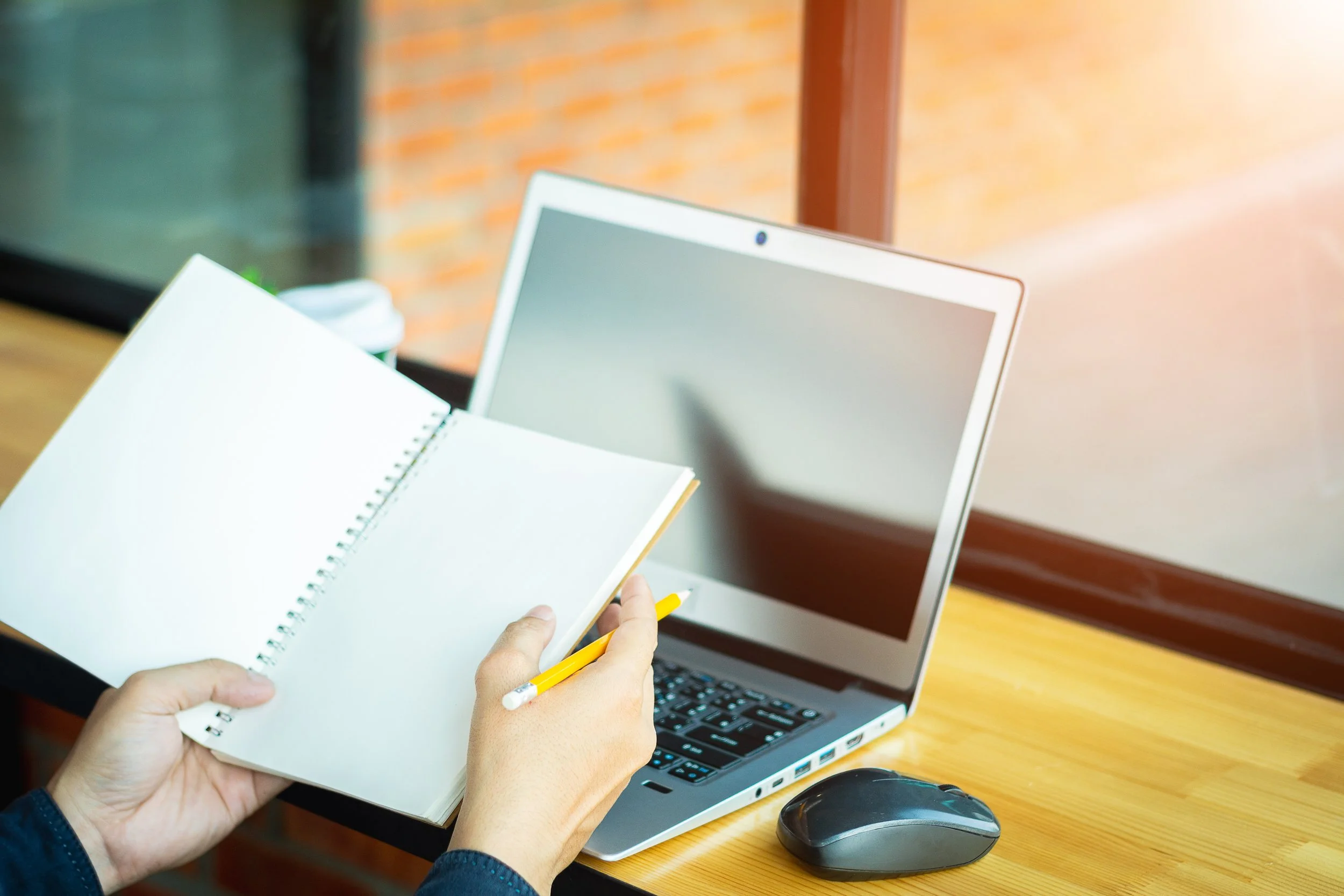 Person holding open notebook and yellow pencil at wooden desk with open laptop, mouse, and coffee cup near window with brick wall in background.
