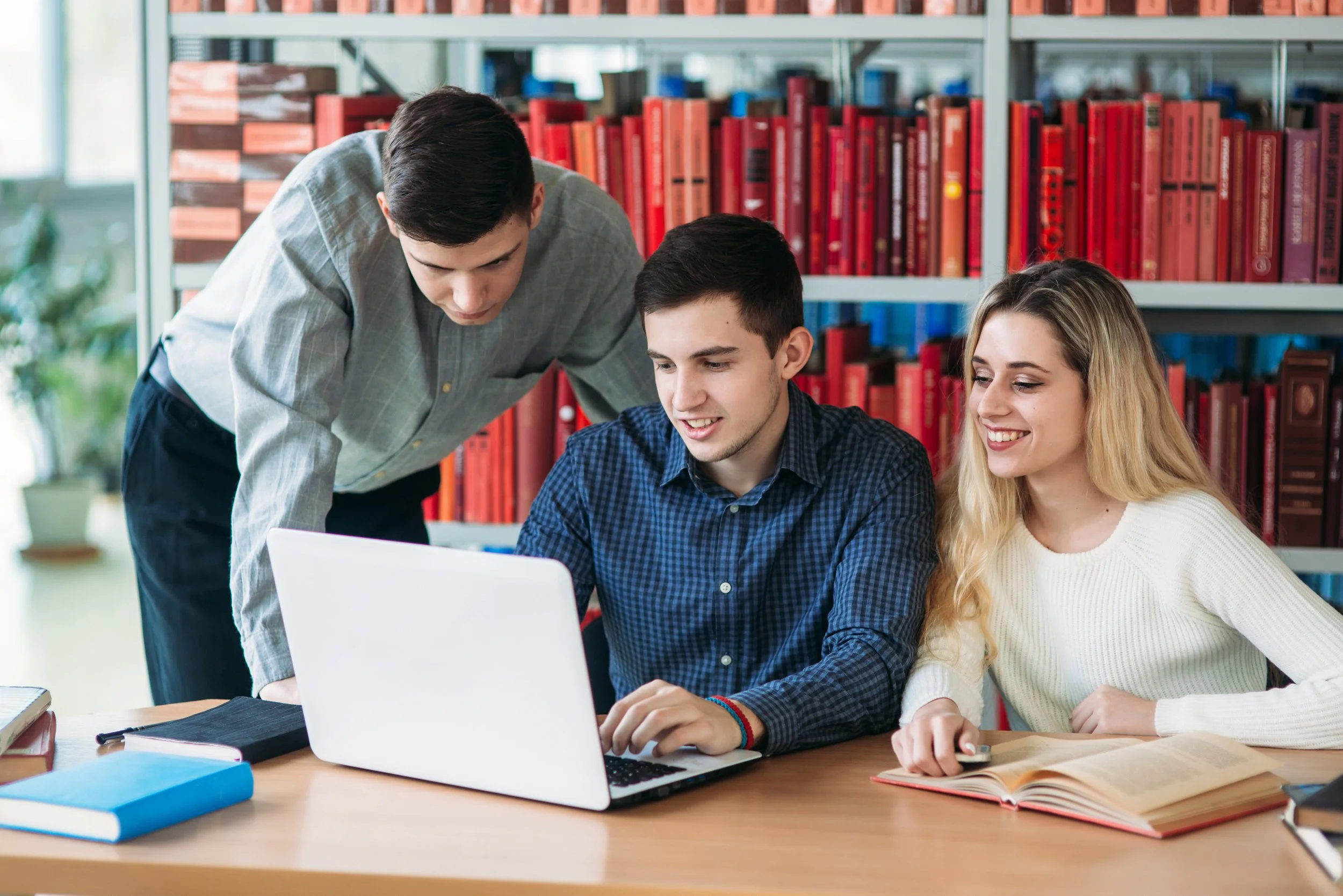 Three young people studying together at a library, looking at a laptop and books.