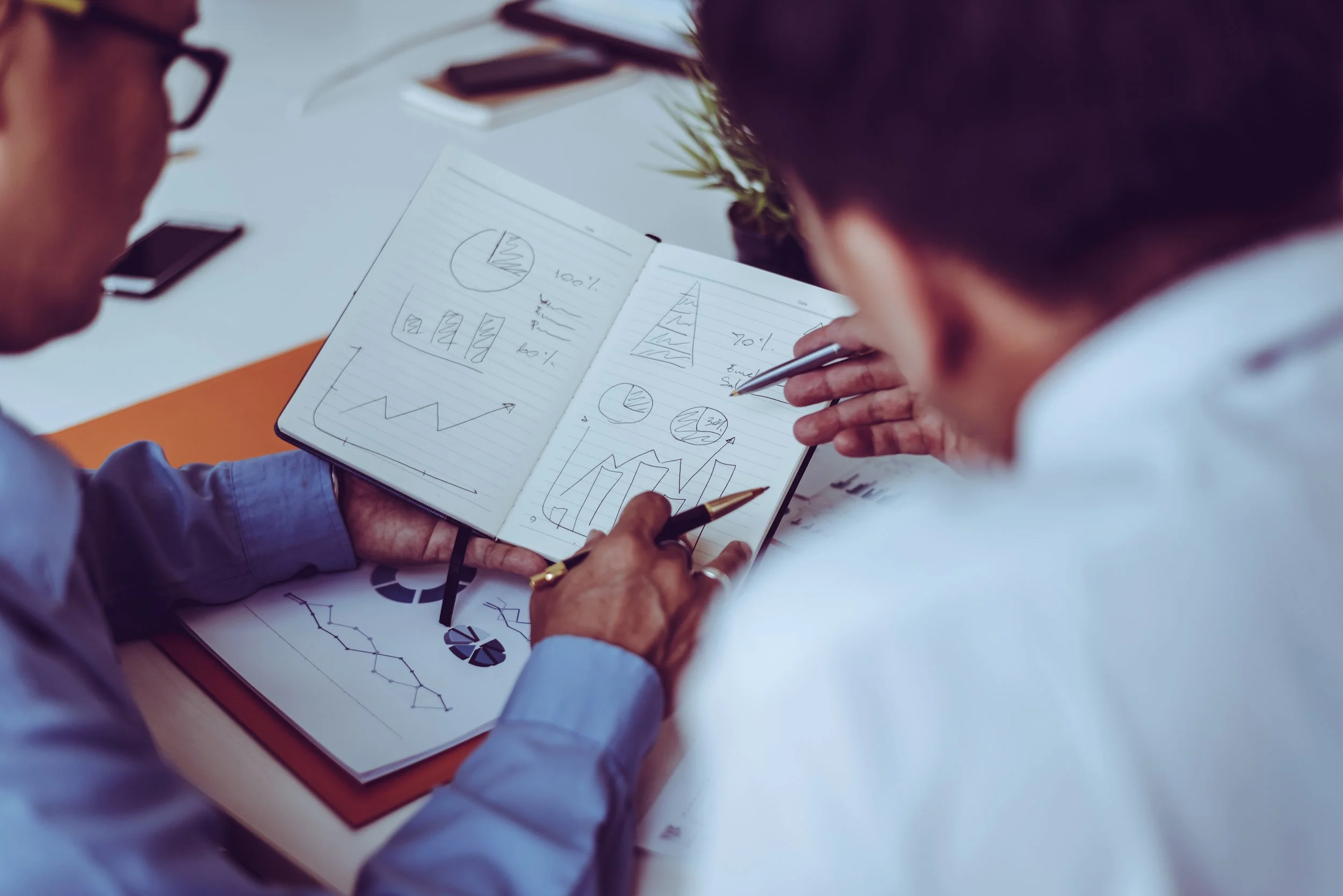 Two people are analyzing and discussing various charts and graphs, including pie charts, bar graphs, and line plots, in a notebook and on printed papers, during a business meeting or presentation.