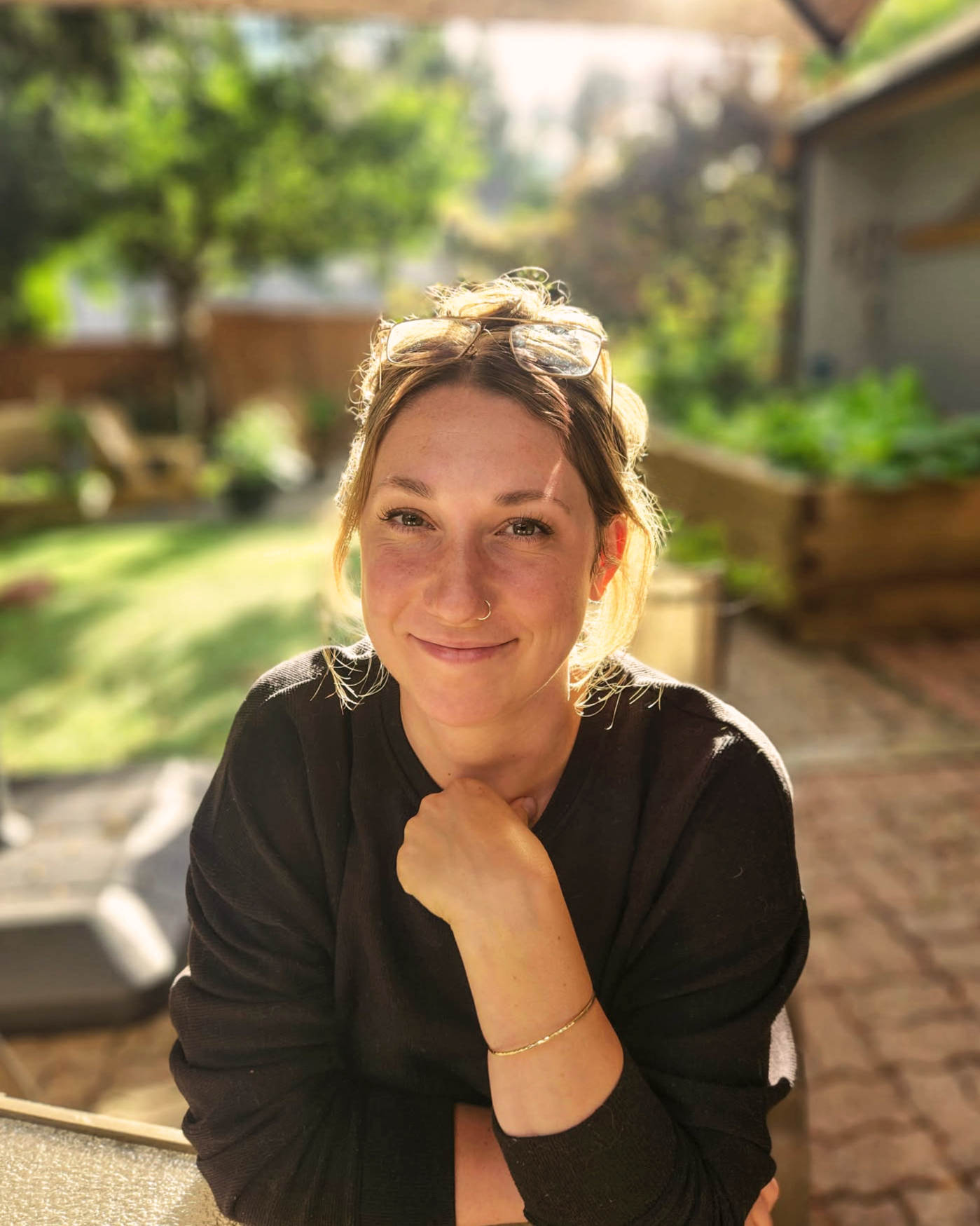 A smiling woman with glasses on her head, wearing a black shirt, sitting outdoors in a garden with green trees and wooden structures in the background, during the daytime.