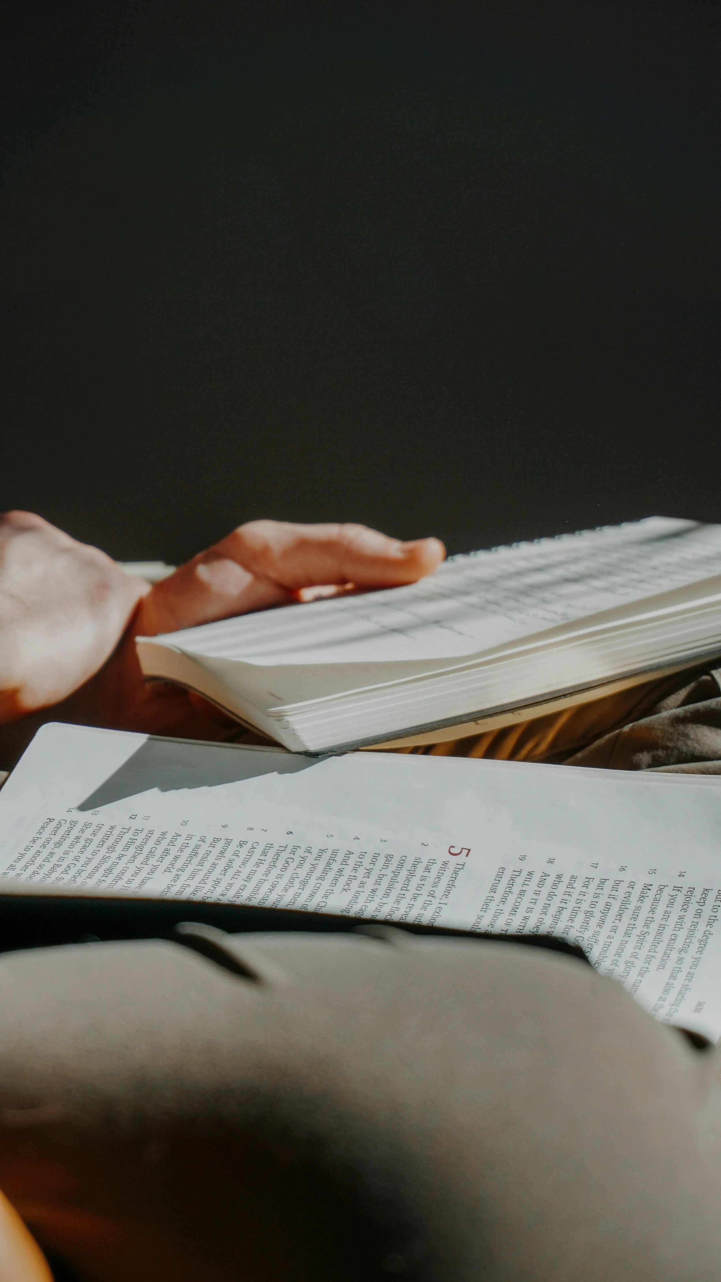 Close-up of a person's hand holding an open book with text, with other papers visible in the foreground and a dark background.