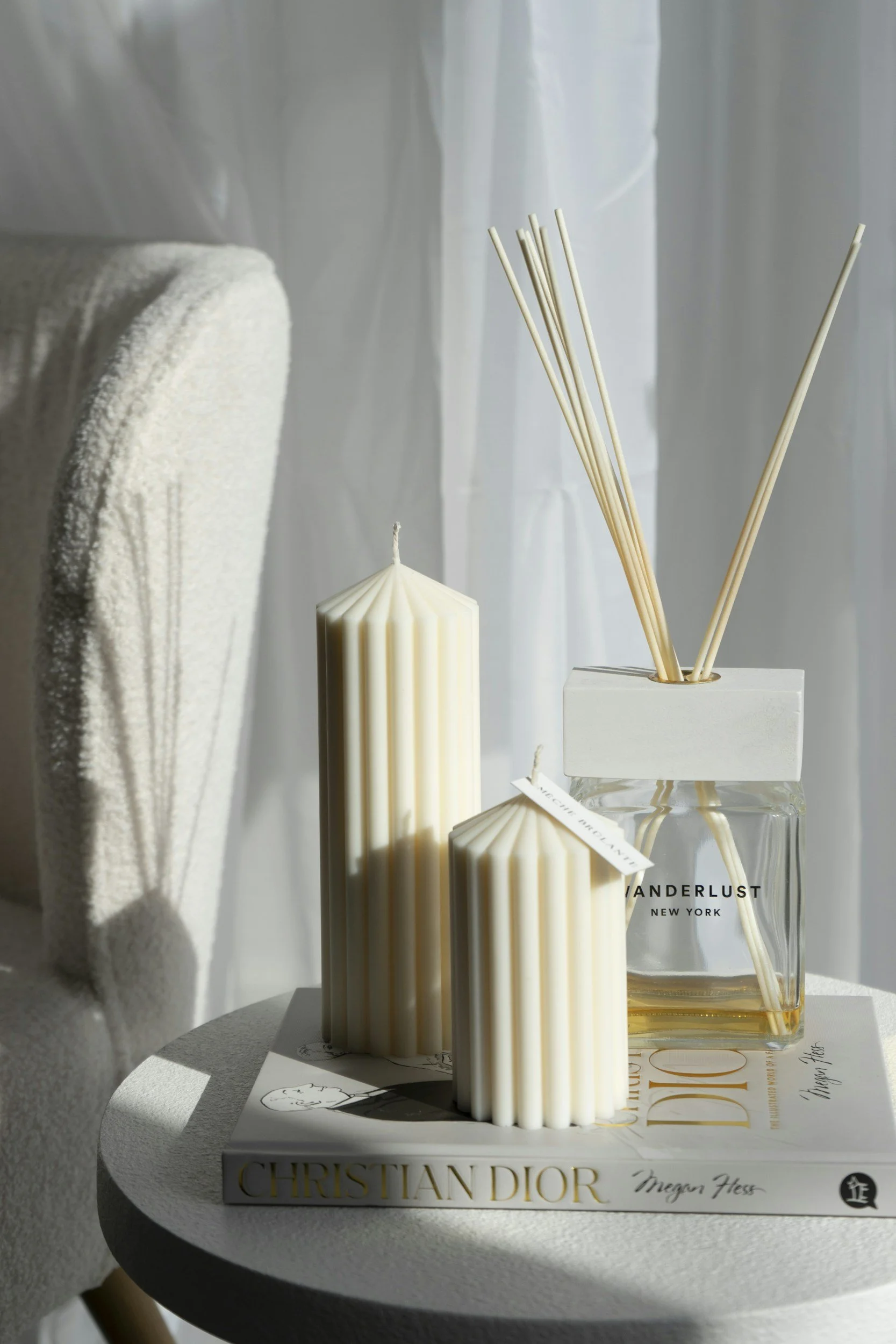 Decorative candles and reed diffuser on a white side table with a fashion magazine underneath, near a soft armchair in natural light.