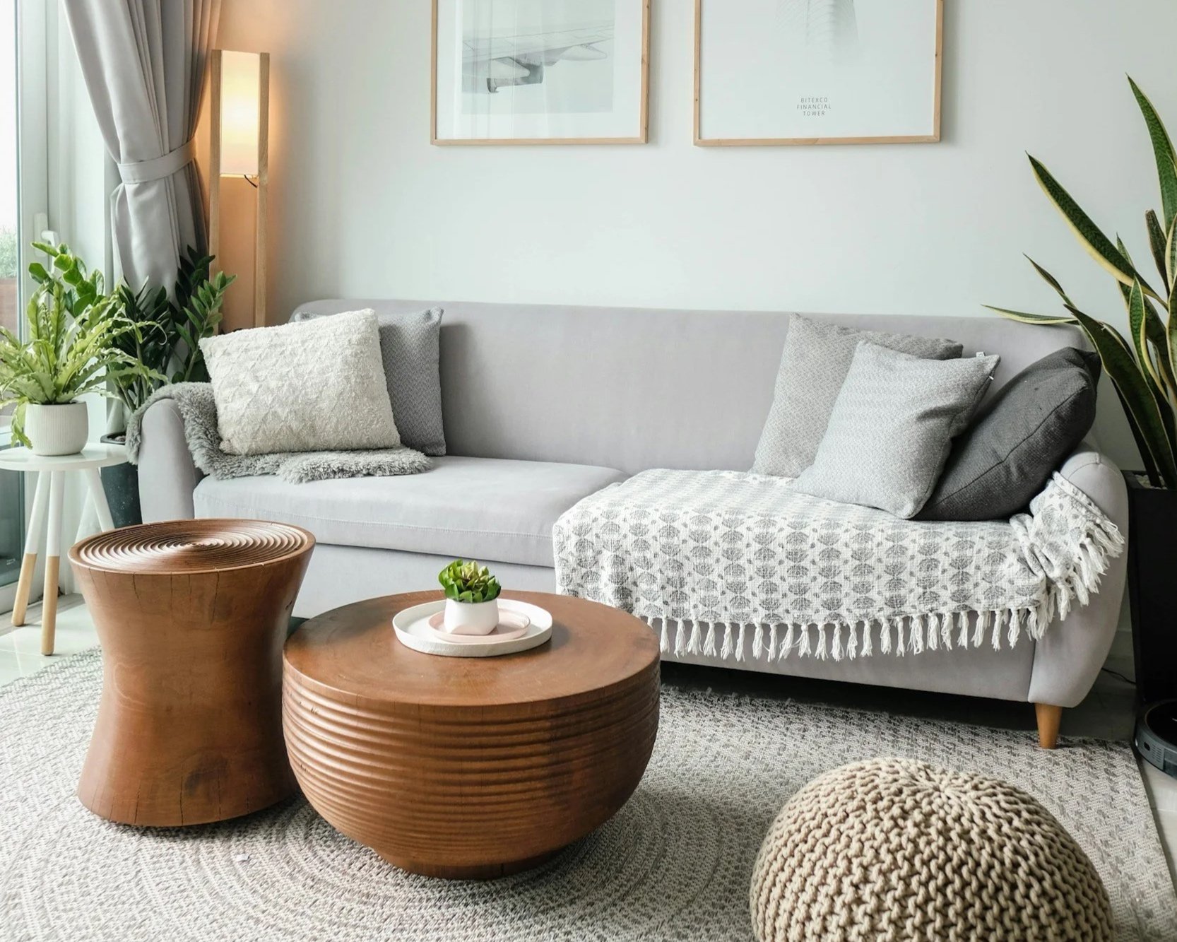 Living room with light gray sofa adorned with various gray and white pillows, a cream and gray throw blanket, surrounded by potted greenery, wooden coffee tables, and framed artwork on the wall.