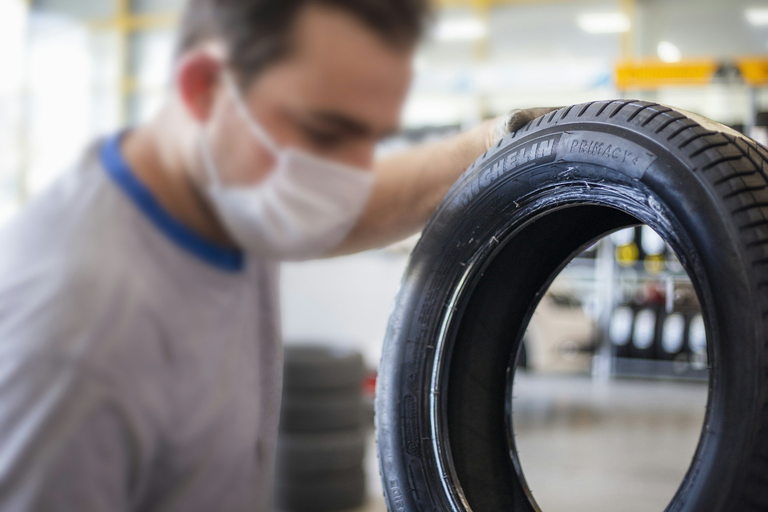 A person wearing a white face mask examines a black Michelin tire in a workshop or tire shop environment.