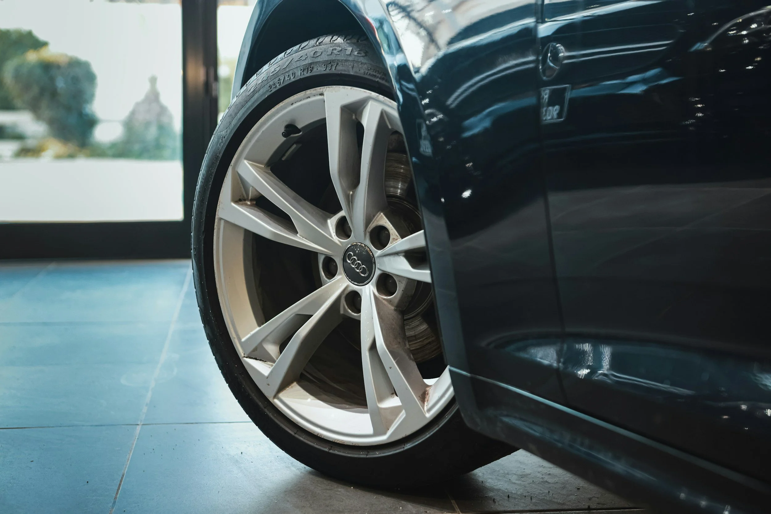 Close-up of a black Audi car wheel with a silver rim, parked inside near a glass door.