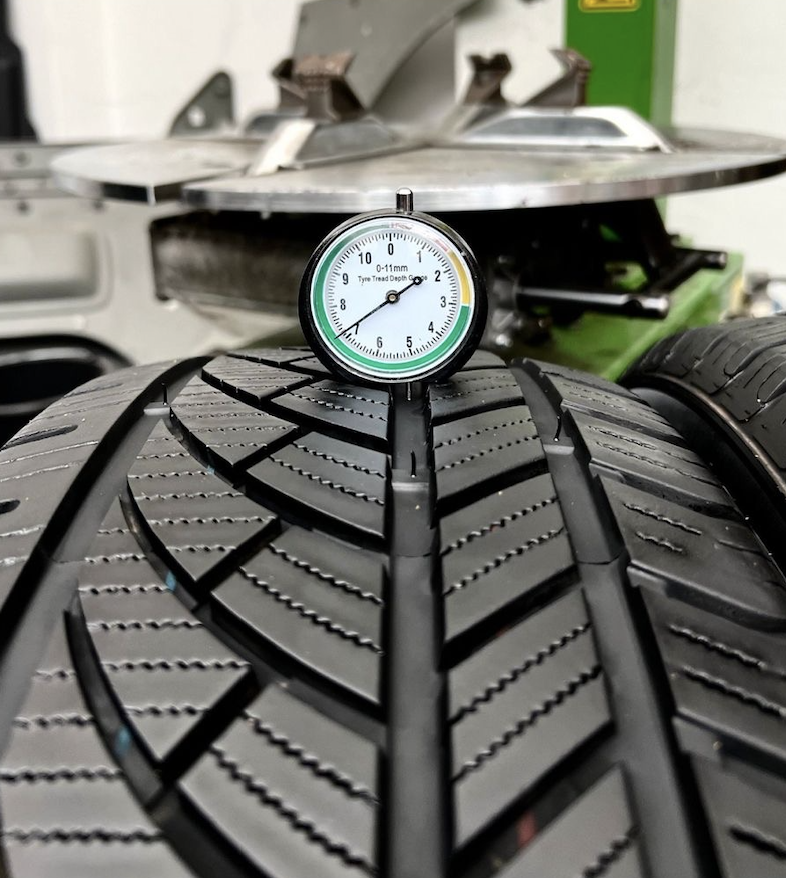 Close-up of a car tire tread depth gauge placed on a tire, with a background showing other tires and machinery in a garage or workshop.
