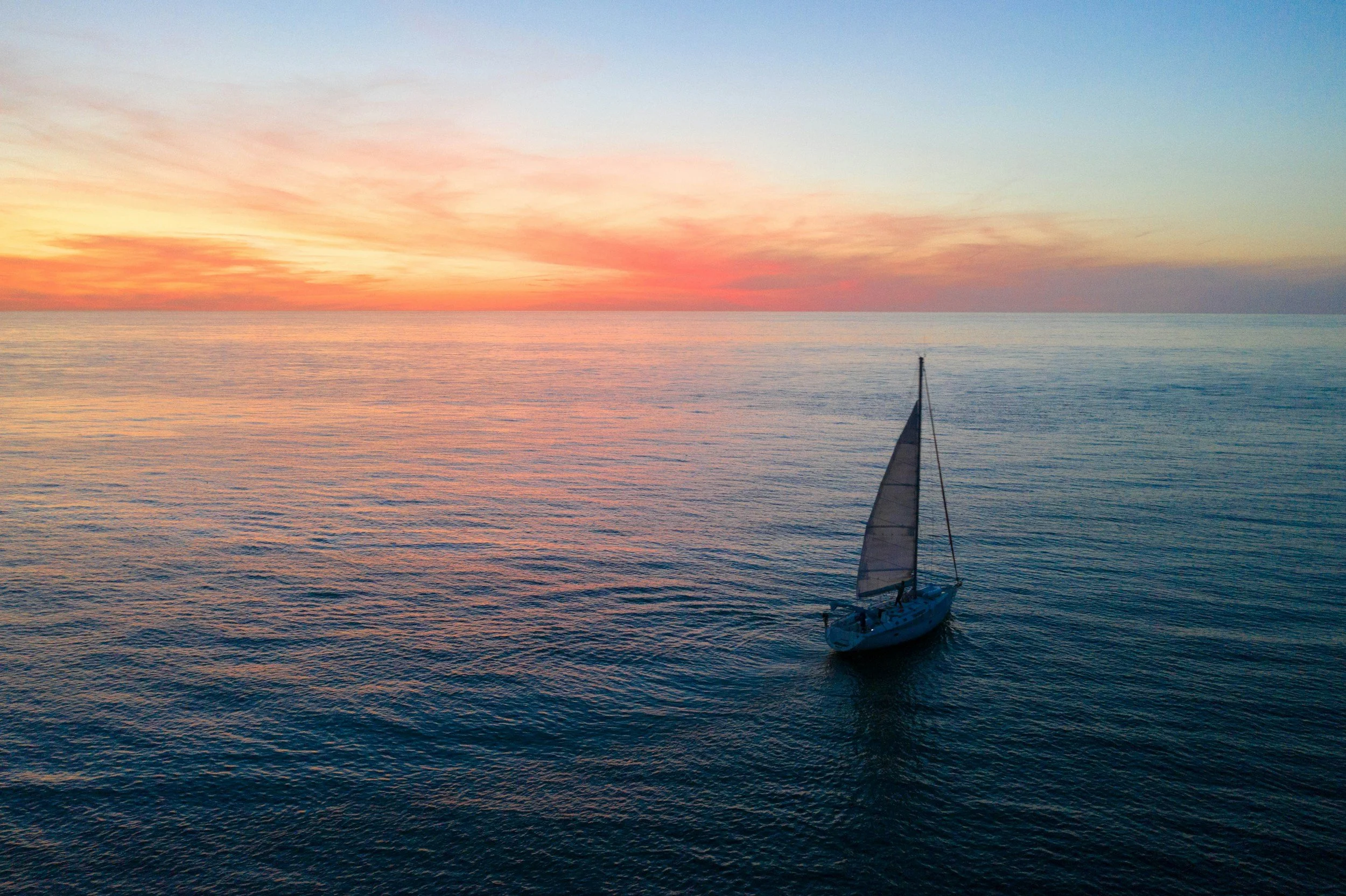 A sailboat floating on calm ocean water at sunset, with a colourful sky of pink, orange, and blue hues.