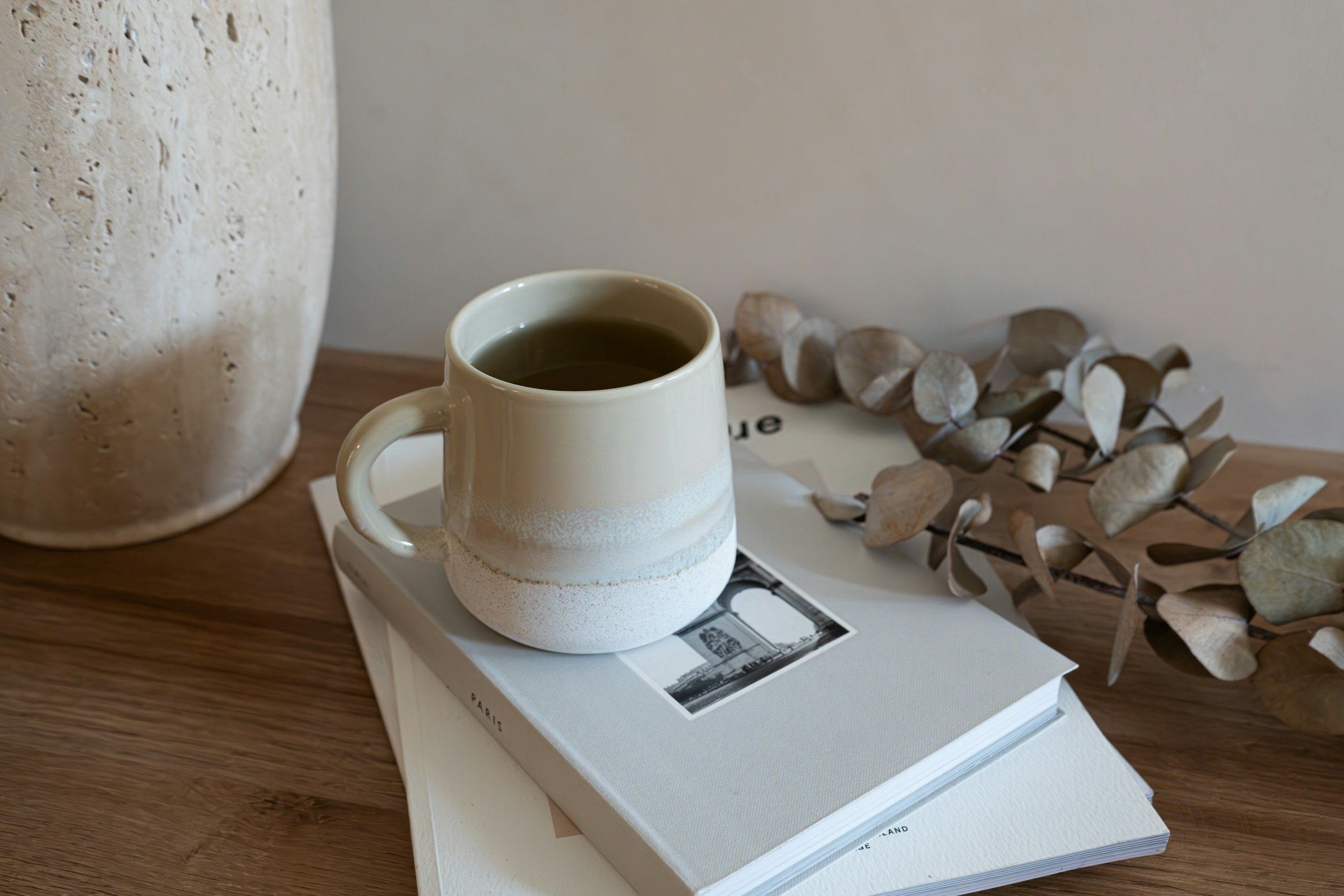 A white ceramic mug with dark coffee, placed on top of a book on a wooden table, next to dried leaves and a large textured vase.
