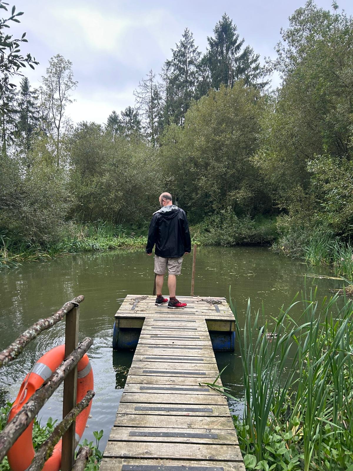 A man standing on a small wooden dock over a narrow body of water, surrounded by dense trees and tall grasses, with an orange life preserver attached to the dock railing.