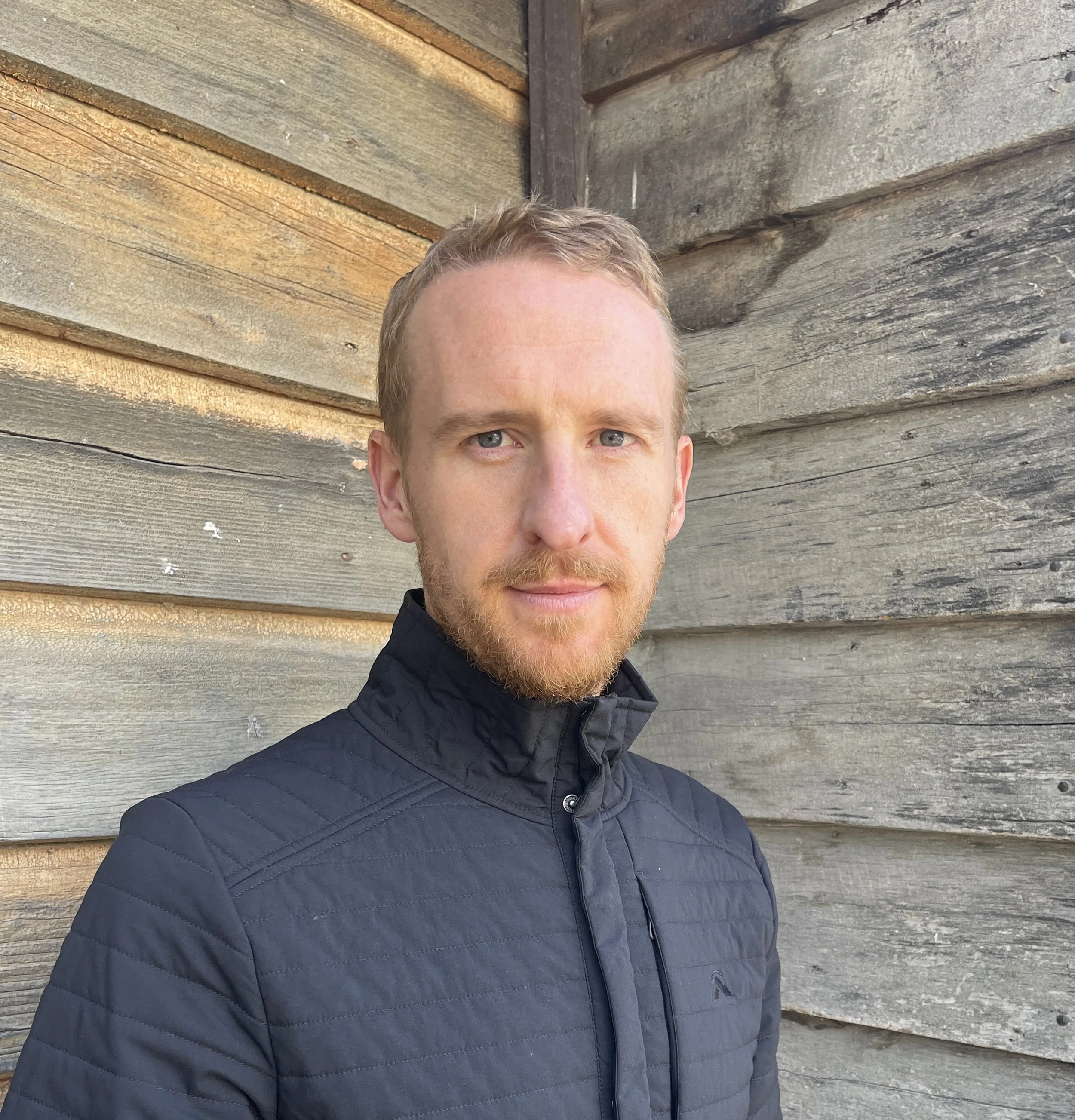 A young man with red hair and a beard taking a selfie outdoors against a weathered wooden wall, wearing a black quilted jacket.