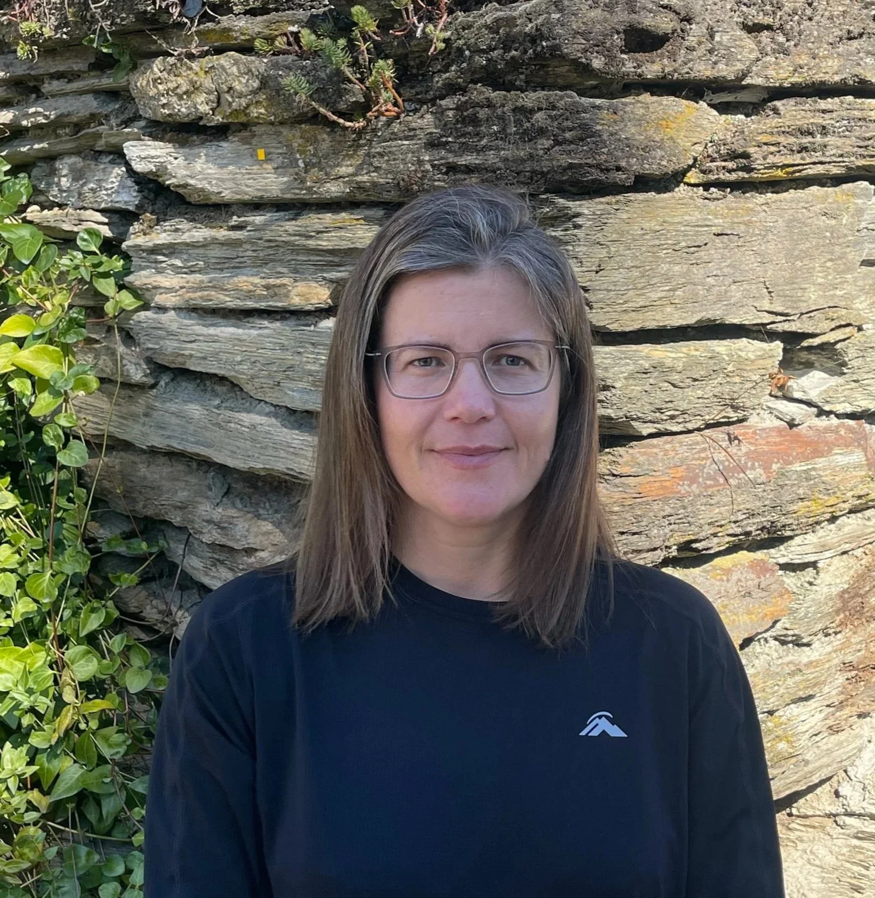 A woman with glasses and long brown hair standing in front of a textured stone wall and green foliage.