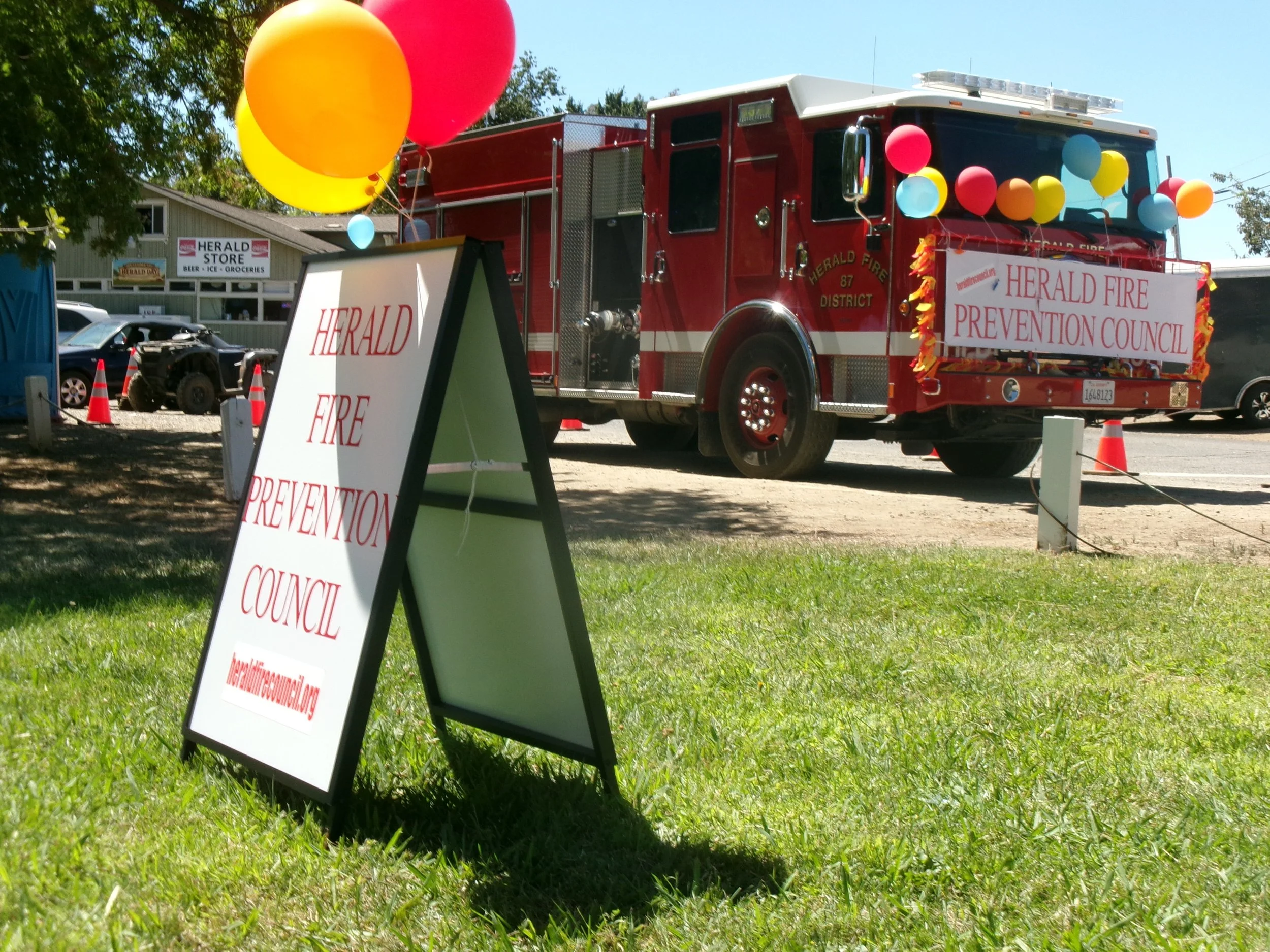 A red fire truck decorated with colorful balloons and two banners for the Herald Fire Prevention Council, parked on a roadside. A sign standing on the grass in front of the truck displays the same organization name.