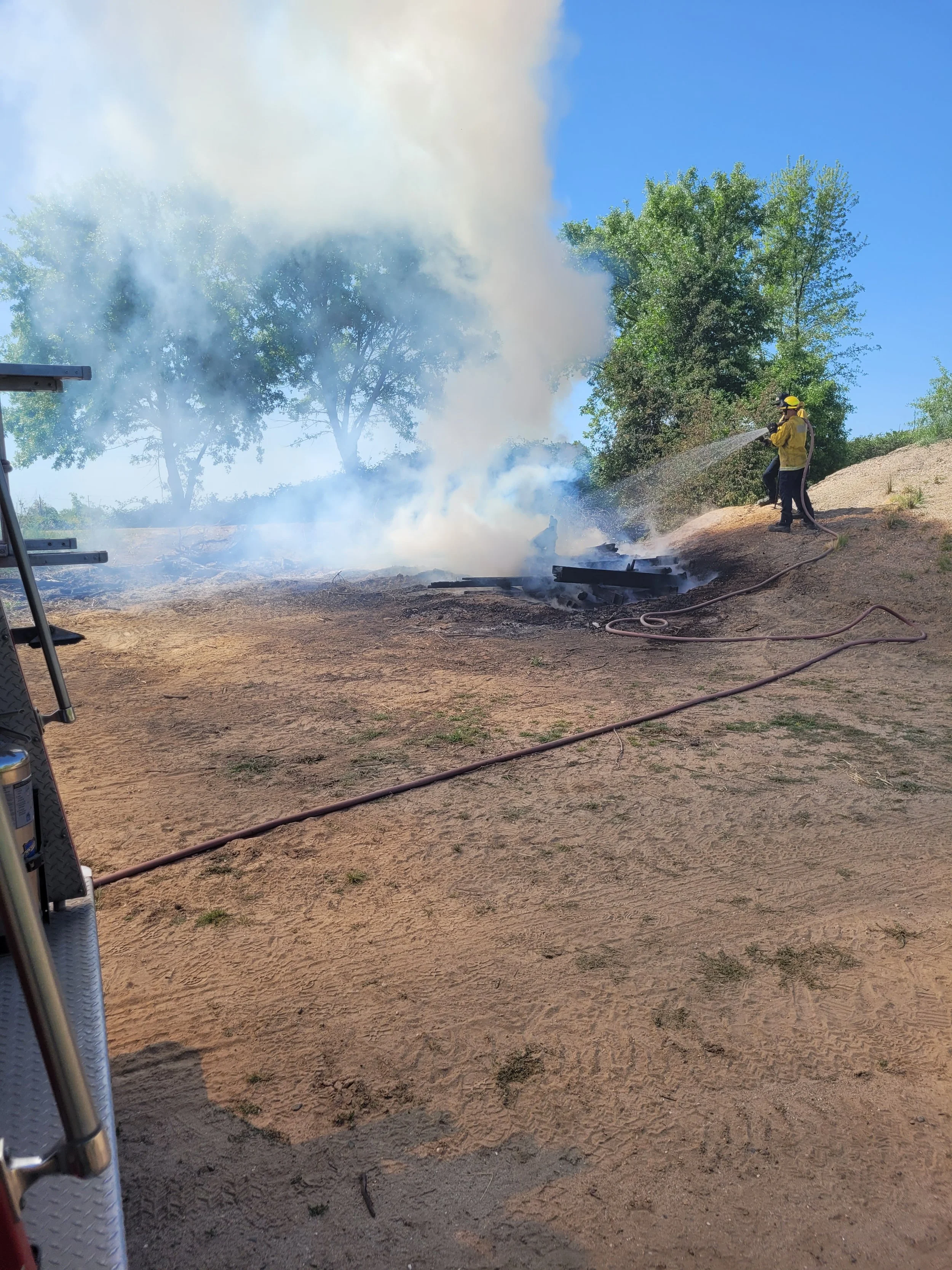 Firefighter spraying water on smoldering debris and smoke in a dry, sandy area with green trees in the background.