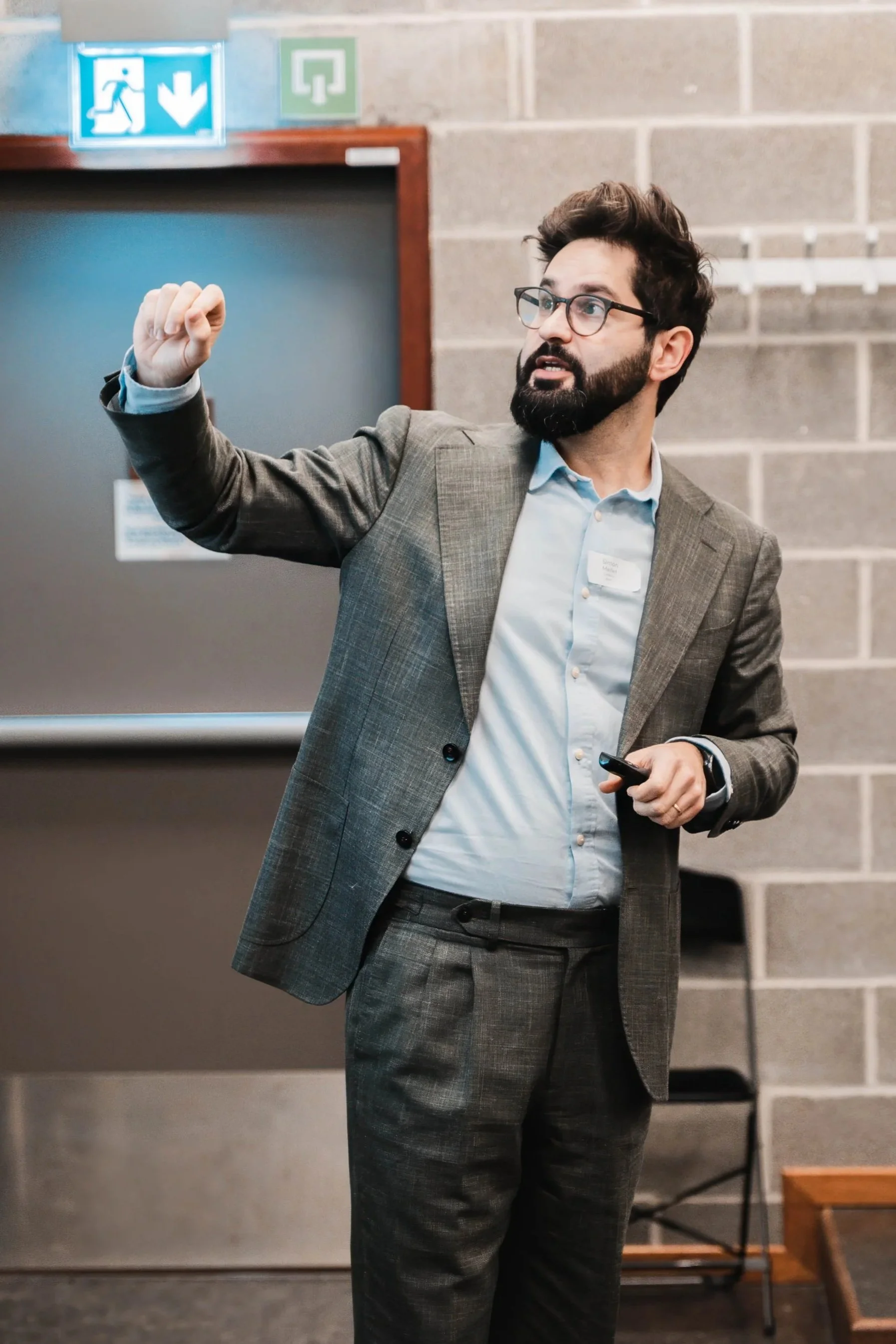 A man in a gray suit and glasses giving a presentation in a conference room, holding a remote control, with emergency exit signs above him.