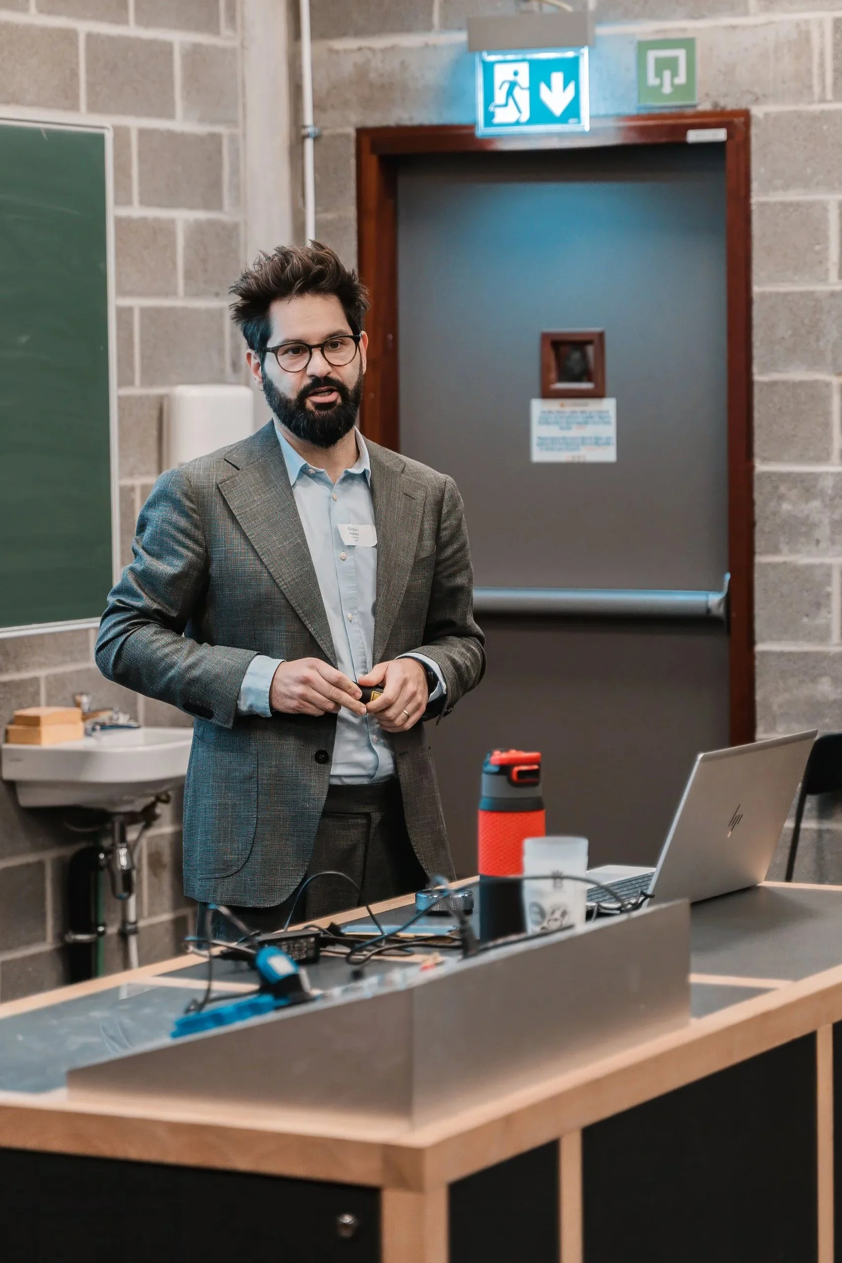 A man with glasses, a beard, and dark hair wearing a brown plaid blazer and light blue shirt, standing at a desk with a laptop, red water bottle, and various cables, in a classroom or conference room with gray brick walls and a green chalkboard.