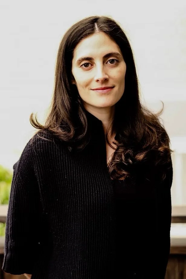 A woman with long, wavy dark hair and light skin, wearing a black sweater, smiling slightly, in an indoor setting with a neutral background.