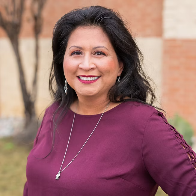 A woman with dark hair wearing a maroon blouse and jewelry smiling outdoors with a brick building and trees in the background.
