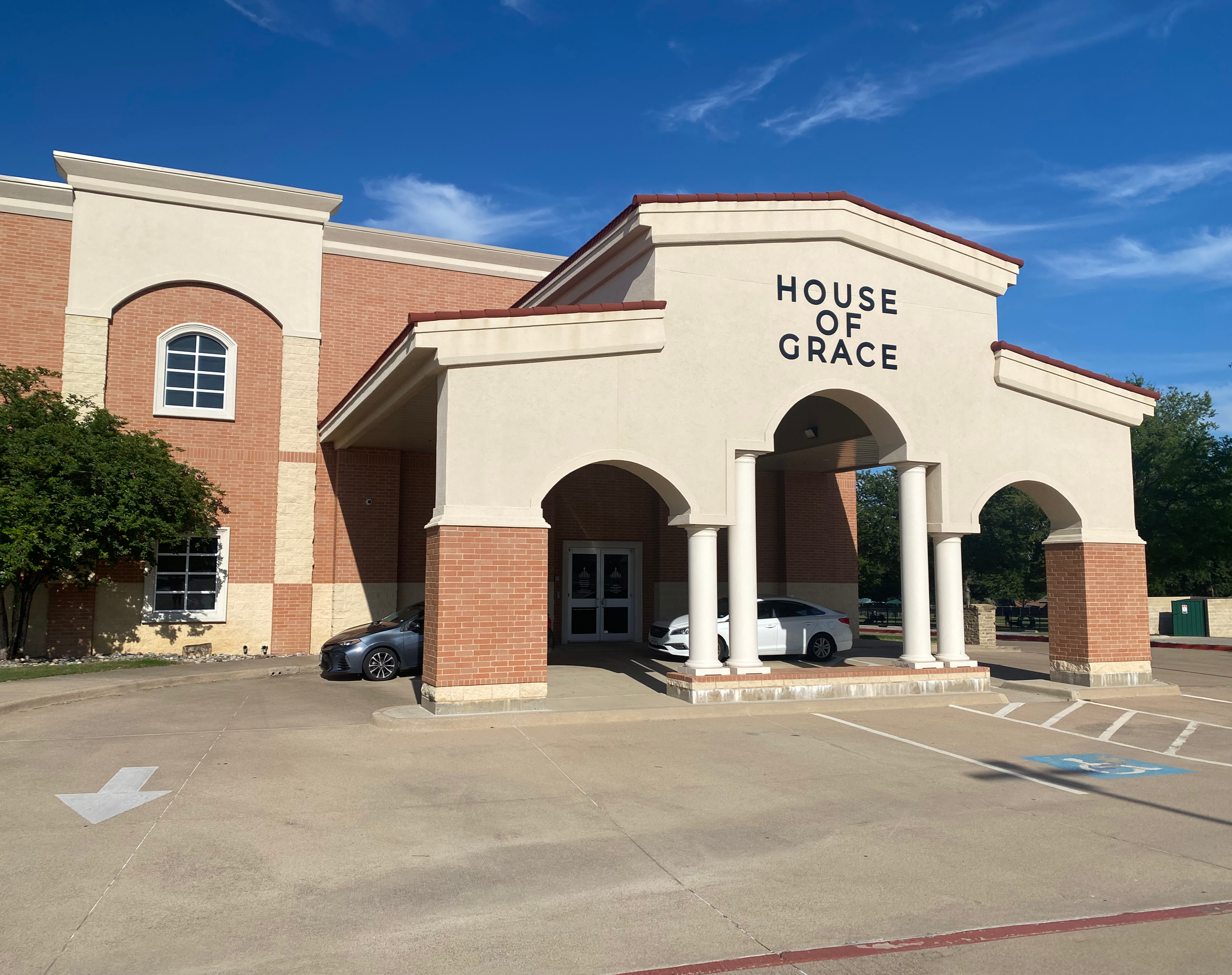 The exterior of a church building named 'House of Grace' with parking spaces and cars in the foreground, under a blue sky with some clouds.