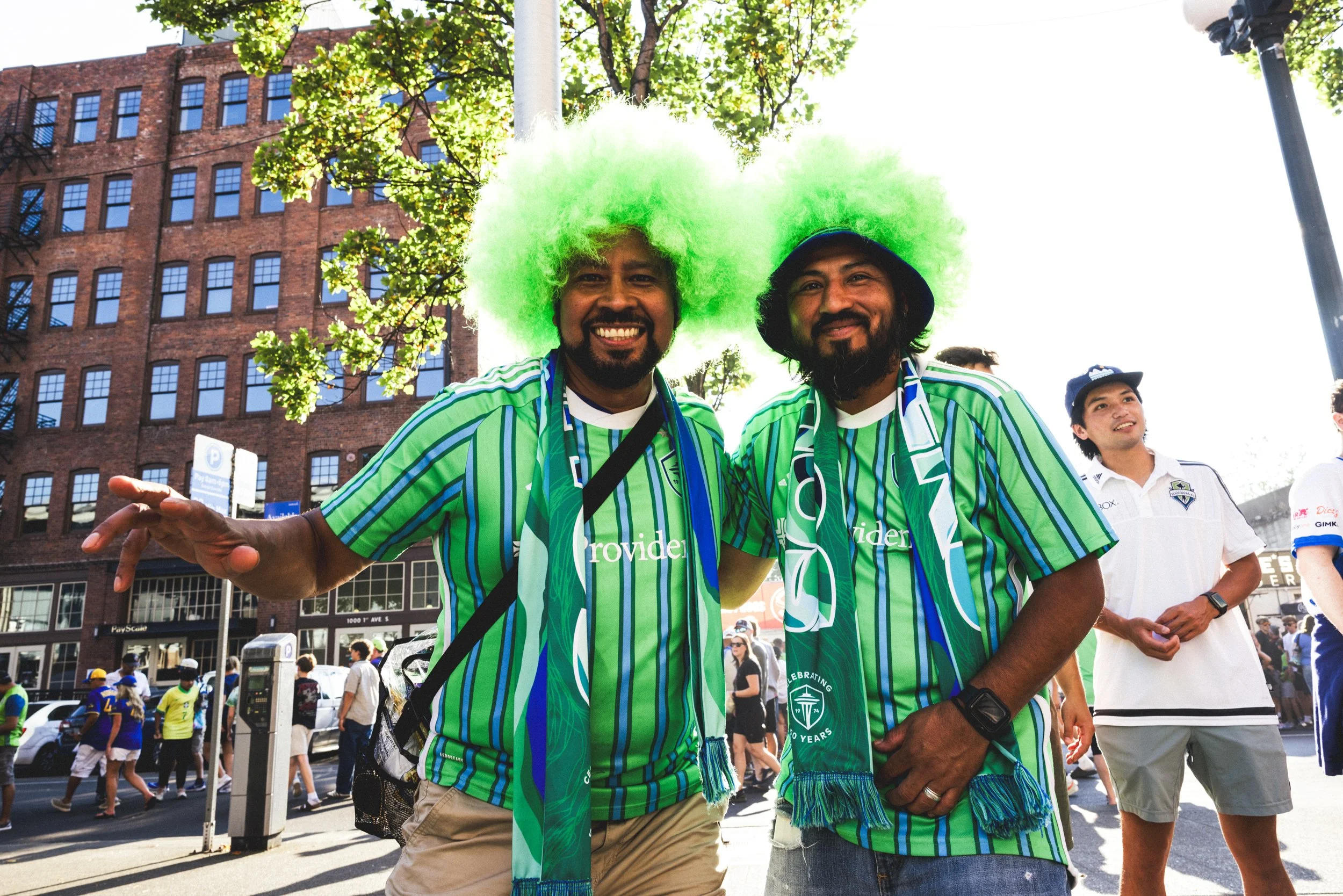 Two smiling men dressed in green and blue Portland Timbers soccer jerseys and green wigs, standing outside on a sunny day at a sports event, with a crowd and brick building in the background.
