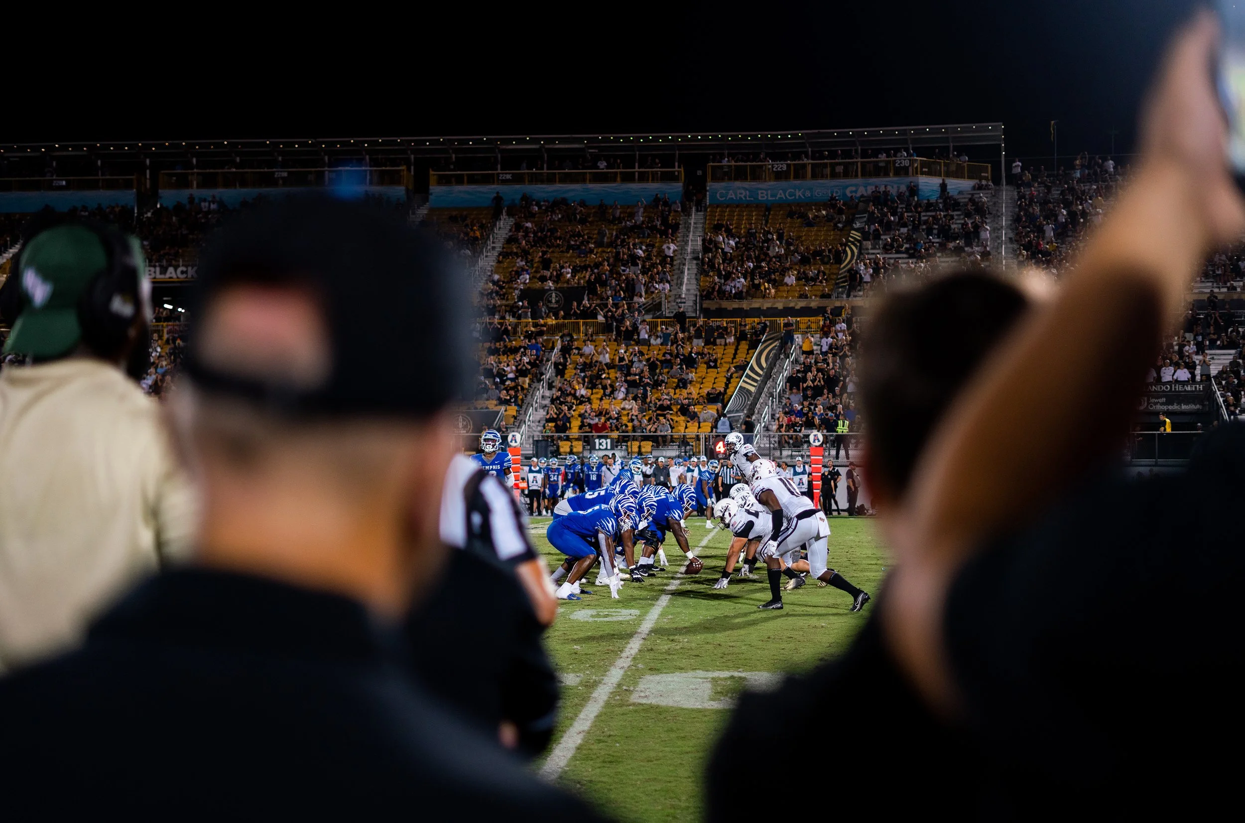 American football game at night, with players in blue and white uniforms lined up on the field, spectators in the stands, and coaches and officials watching from the sideline.