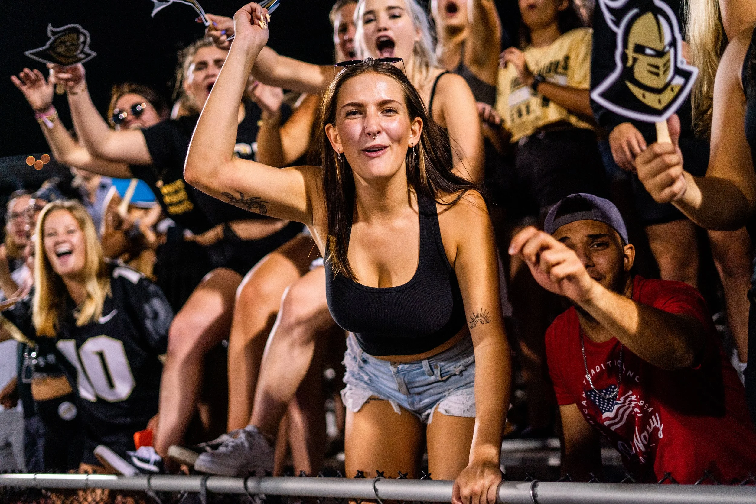 Group of cheering people at a nighttime sports event, with young women and men showing enthusiasm, some holding signs or wearing sports jerseys.