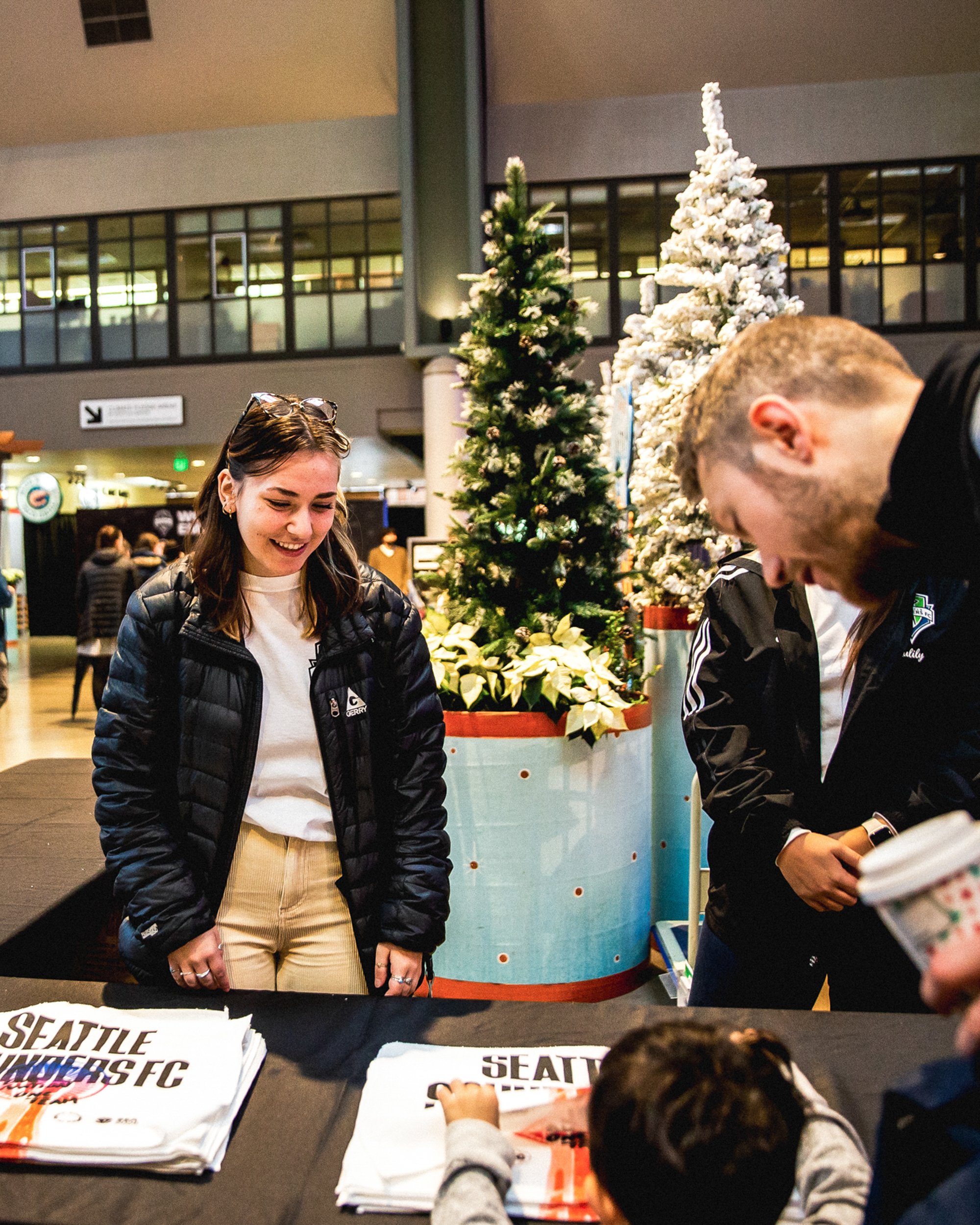 Two people at a table with white Seattle Sounders FC T-shirts, a young girl is smiling and talking to them, in a festive, indoor setting with decorated Christmas trees in the background.