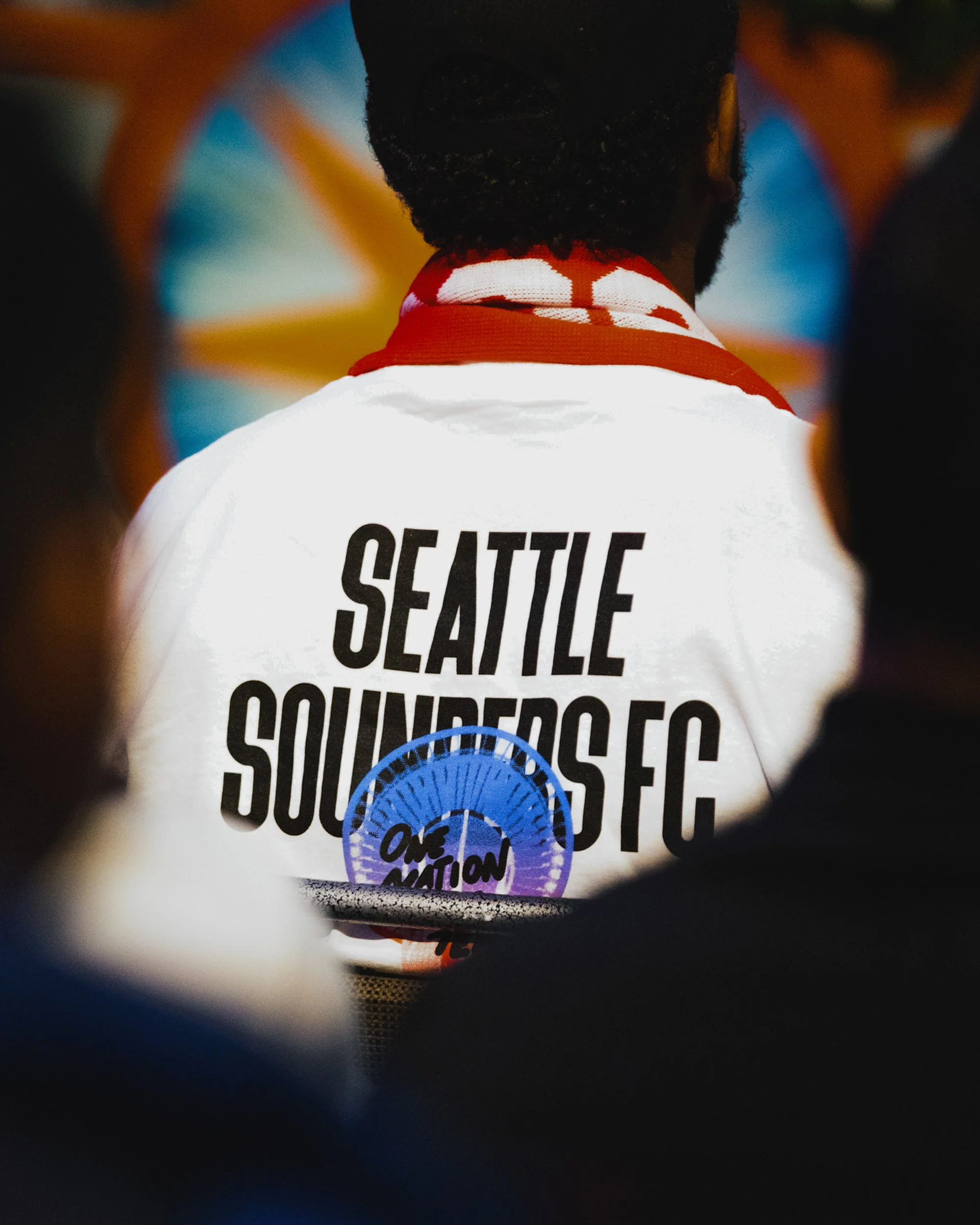 Back of a person wearing a white jacket with 'Seattle Sounders FC' text and a circular logo that says 'One Passion,' attending a sports event.
